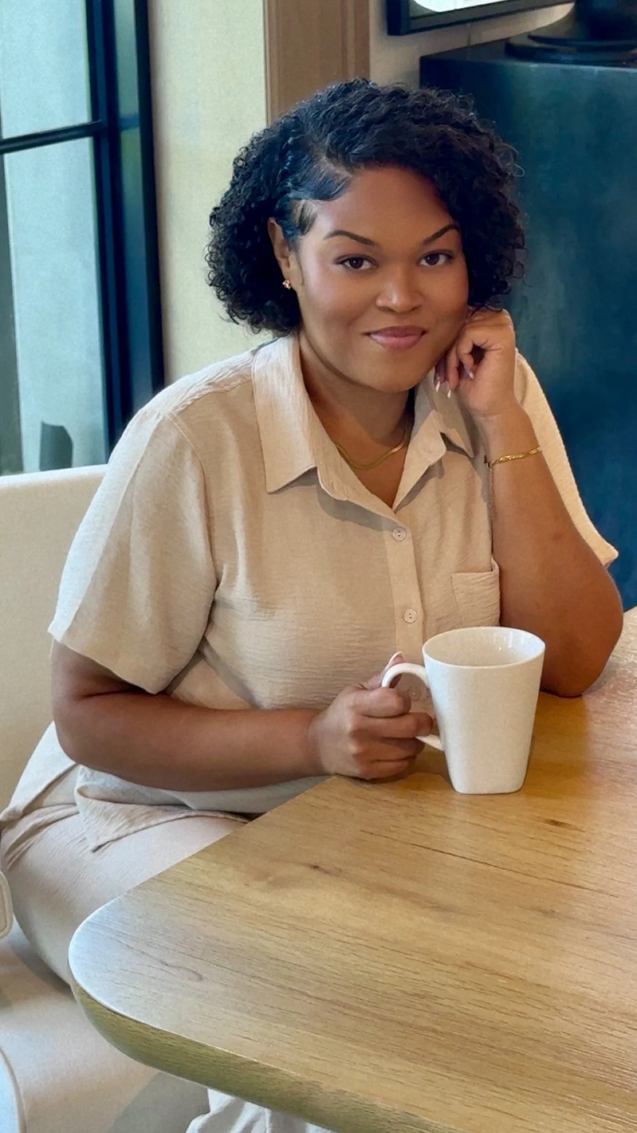 A woman with curly black hair, wearing a beige shirt, sitting at a wooden table, holding a white mug, and resting her chin on her hand, in a cozy indoor setting.