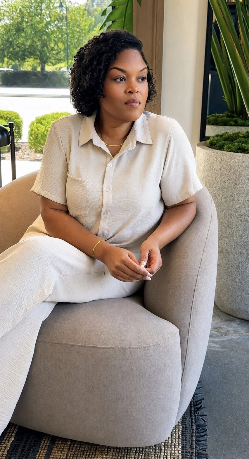 A woman with curly black hair sitting on a beige chair indoors near a large window with greenery outside.