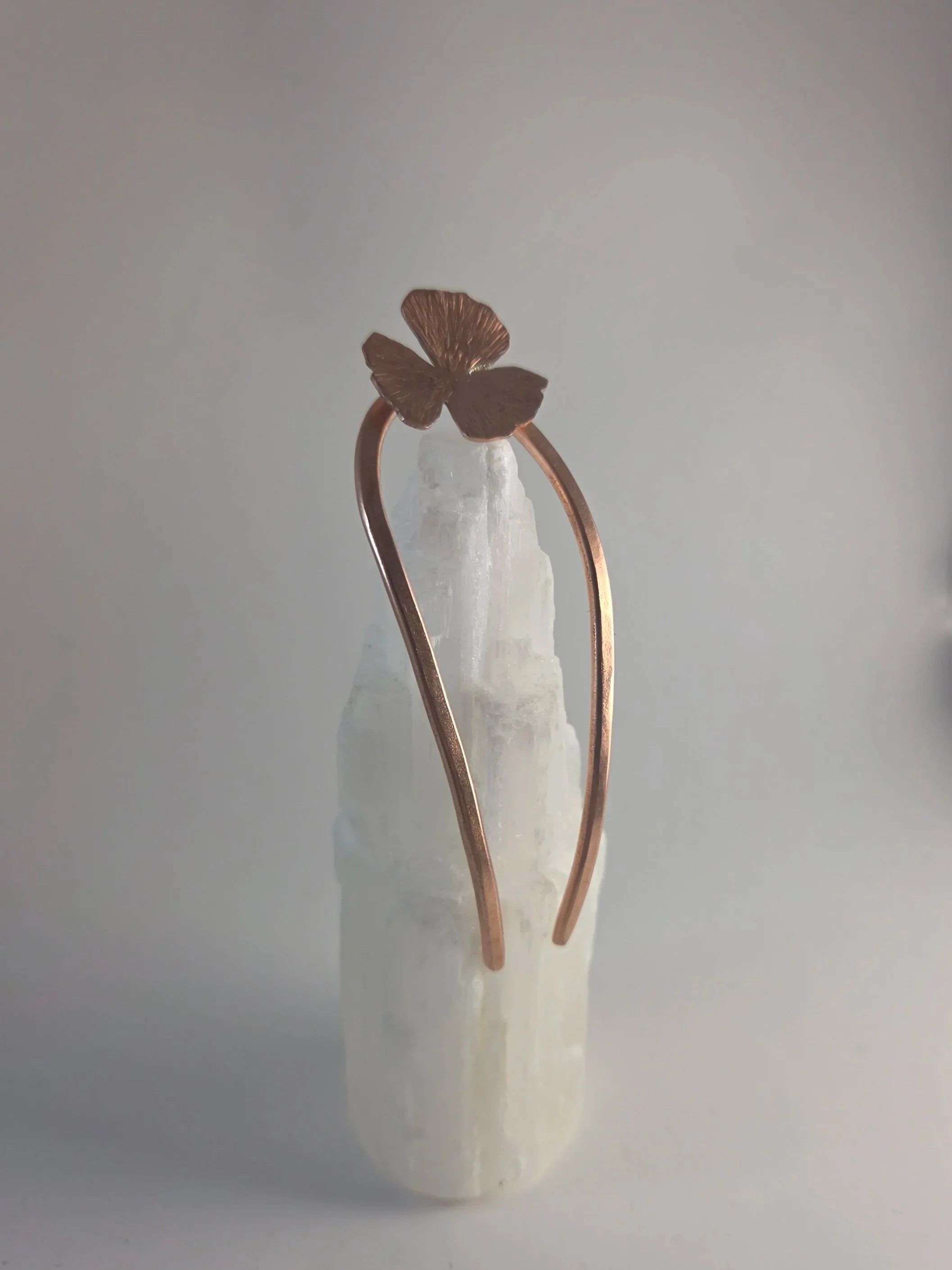 A small bronze butterfly-shaped decorative piece with detailed wings, mounted on a white, textured cone-shaped base, and held by two bent, copper-colored wires.