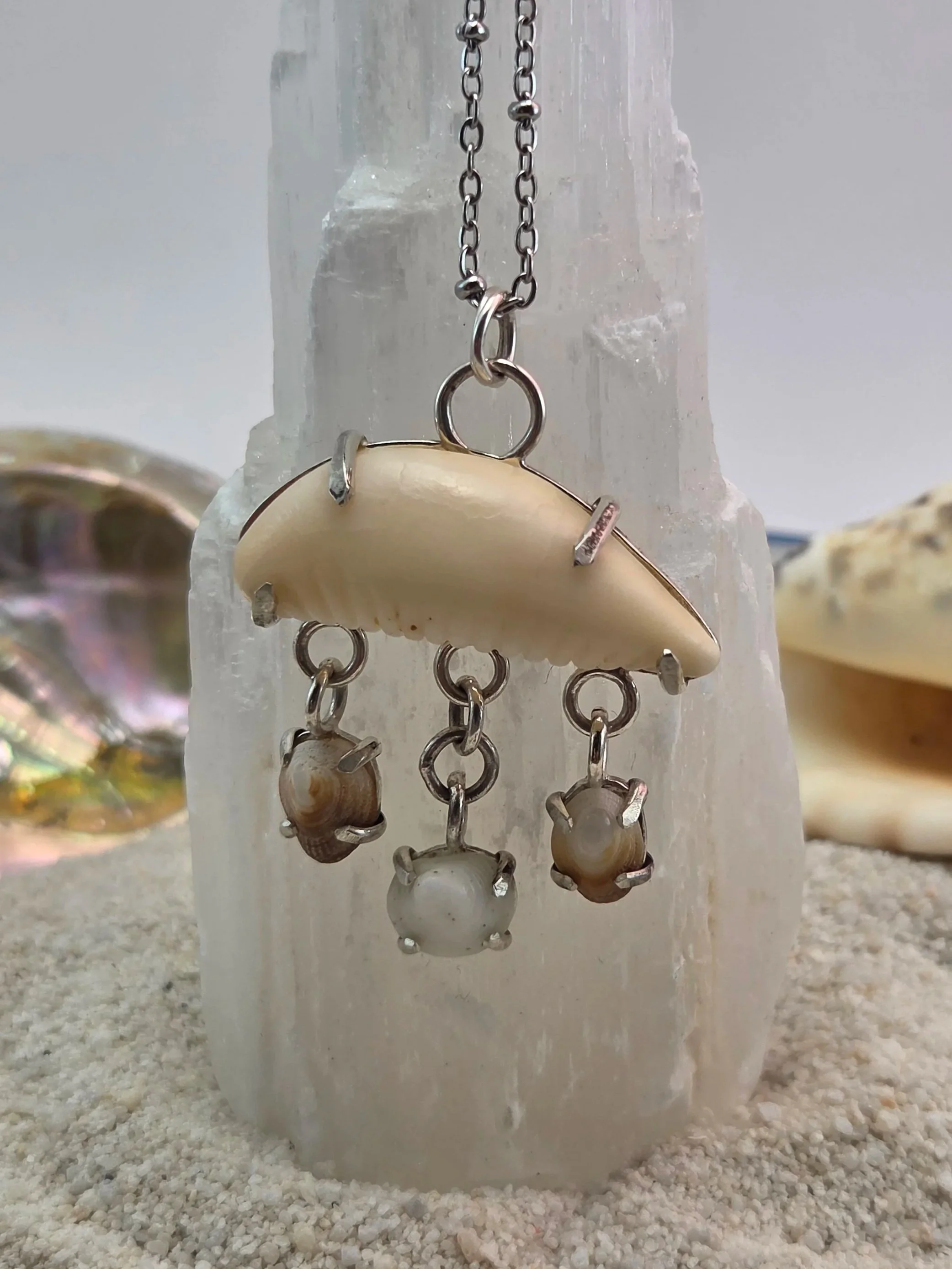 A piece of jewelry featuring a large seashell with smaller shells hanging from it, attached to a silver chain, displayed on a white crystal or mineral formation.