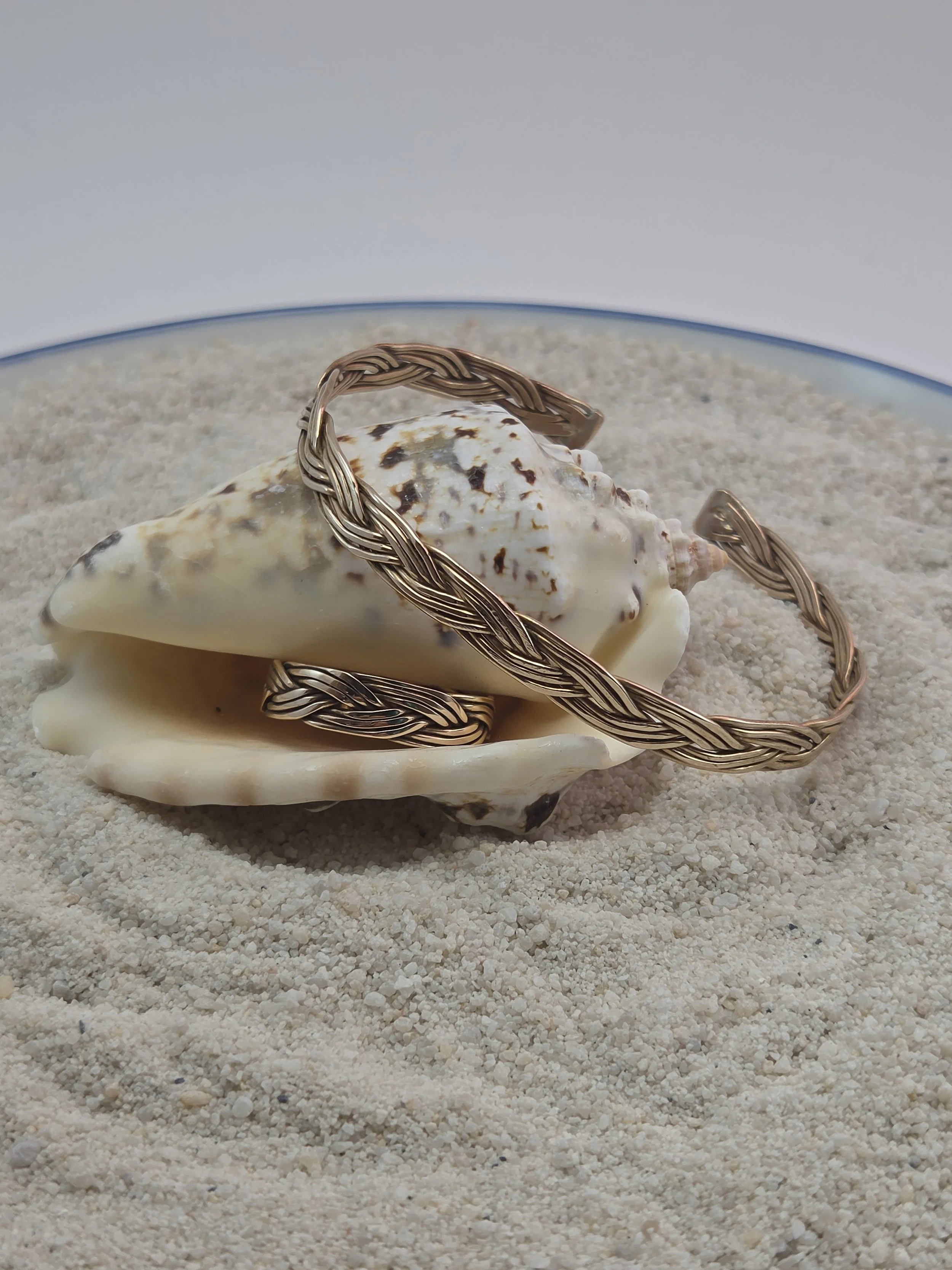 A bronze braided bracelet resting on a large seashell in a glass container filled with sand.
