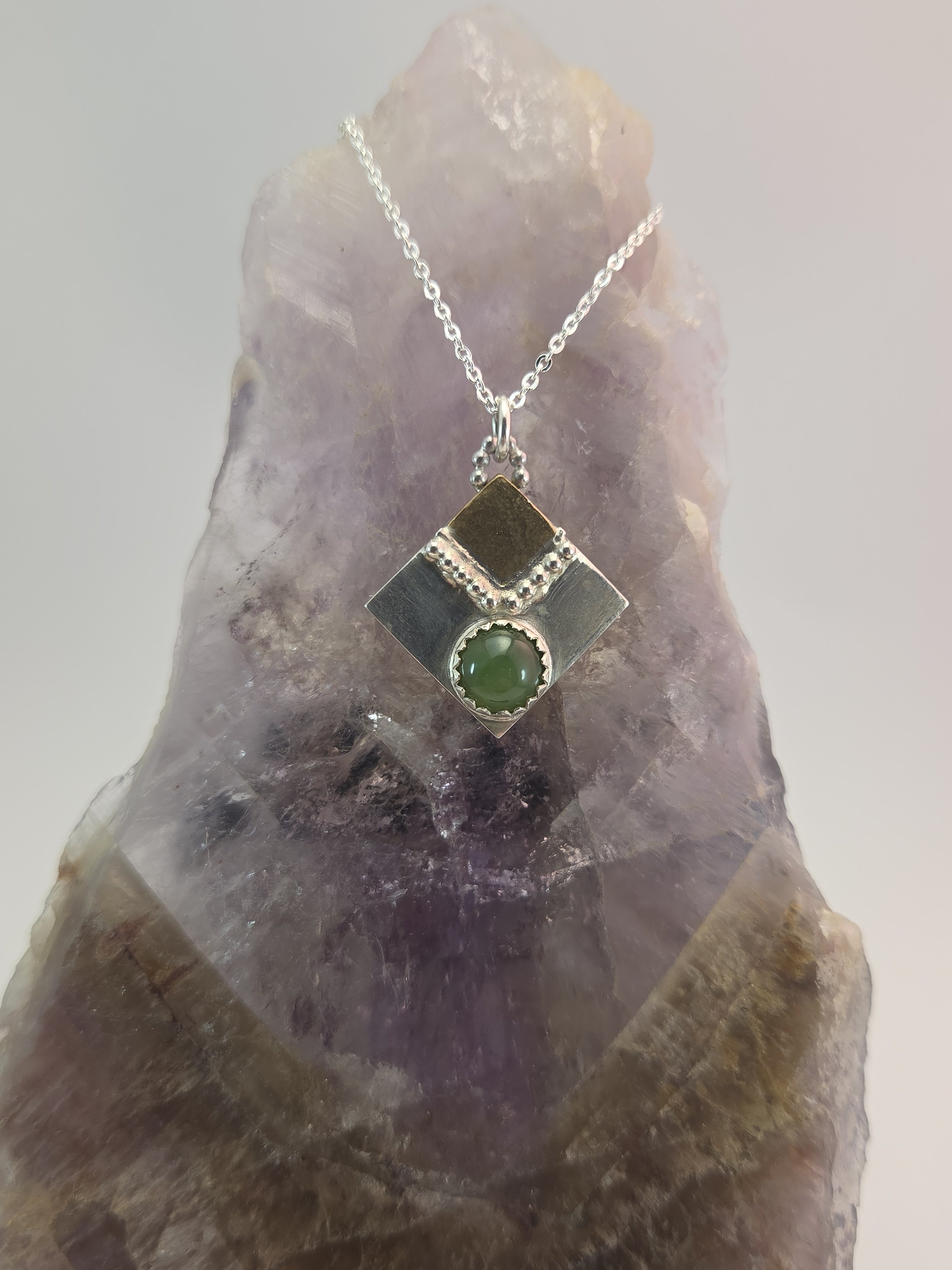 Silver necklace with a square pendant featuring a green gemstone, displayed on a textured purple and brown rock.