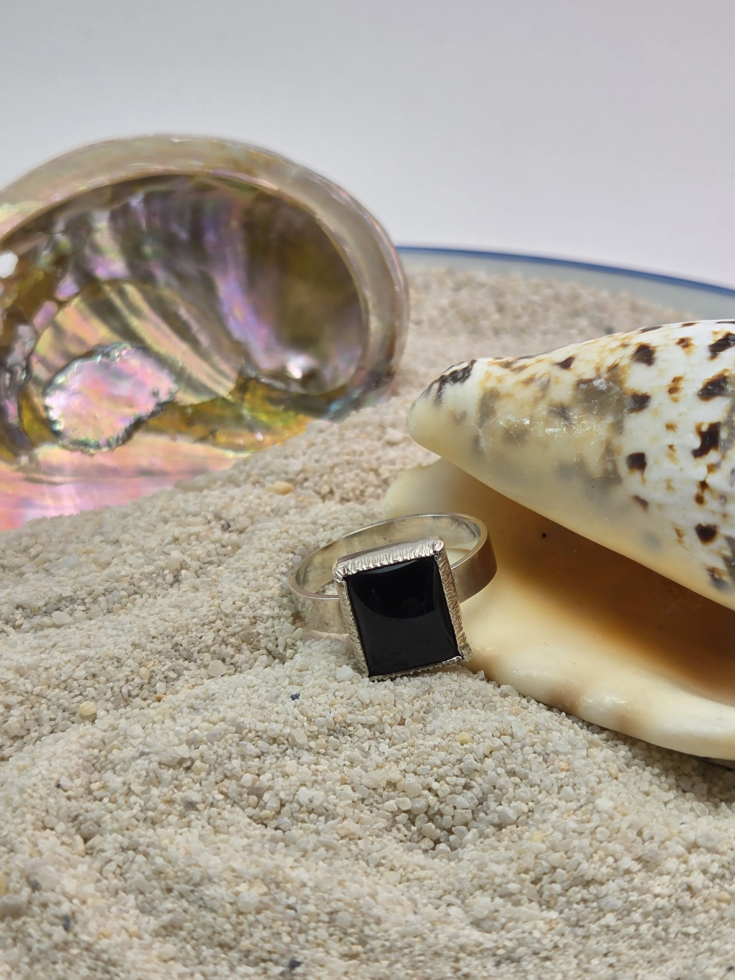 A small black ring with a rectangular black gemstone sitting on white sand, with a large shell and a reflective, iridescent object in the background.