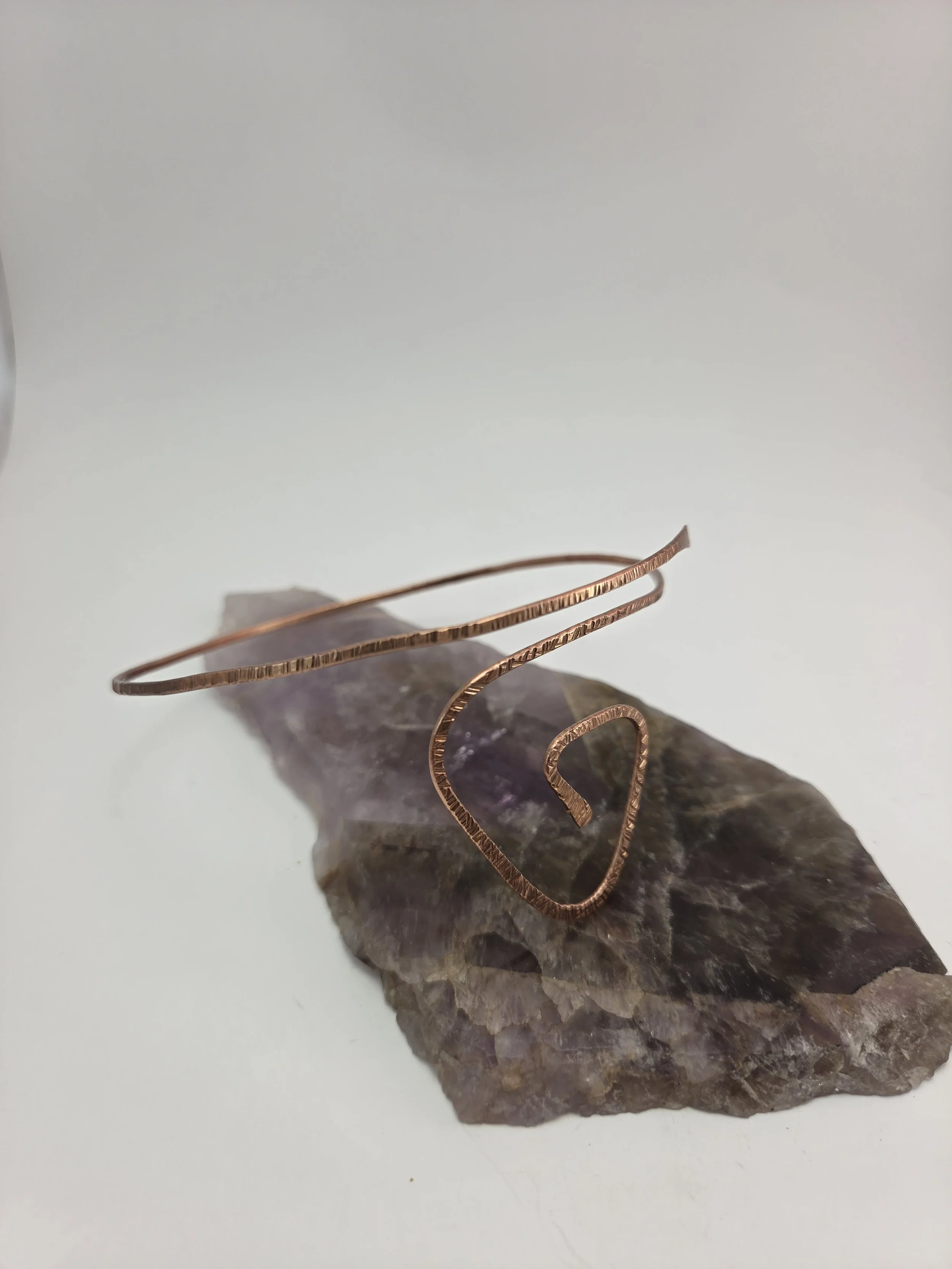 A copper wire arm cuff with a geometric shape resting on a rough purple and gray mineral rock on a white surface.