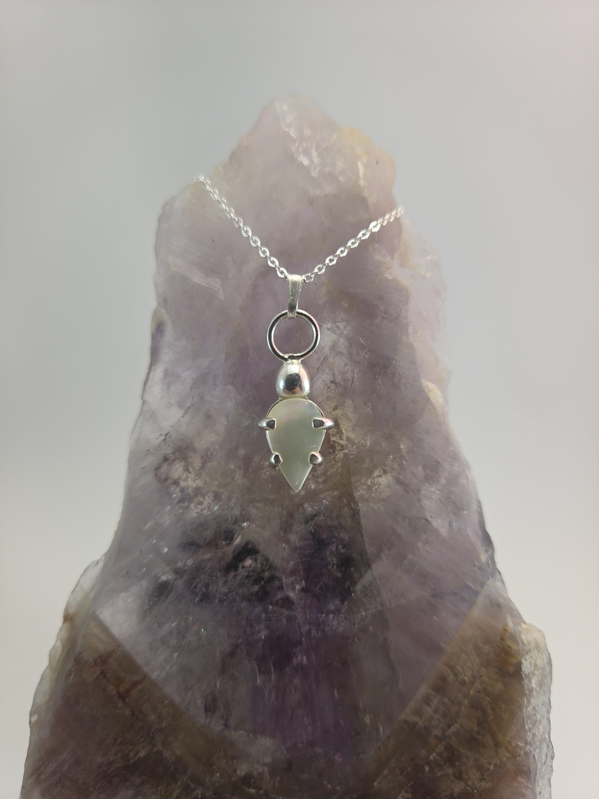 A silver pendant necklace with a clear mother of pearl pendant displayed on a large amethyst geode.