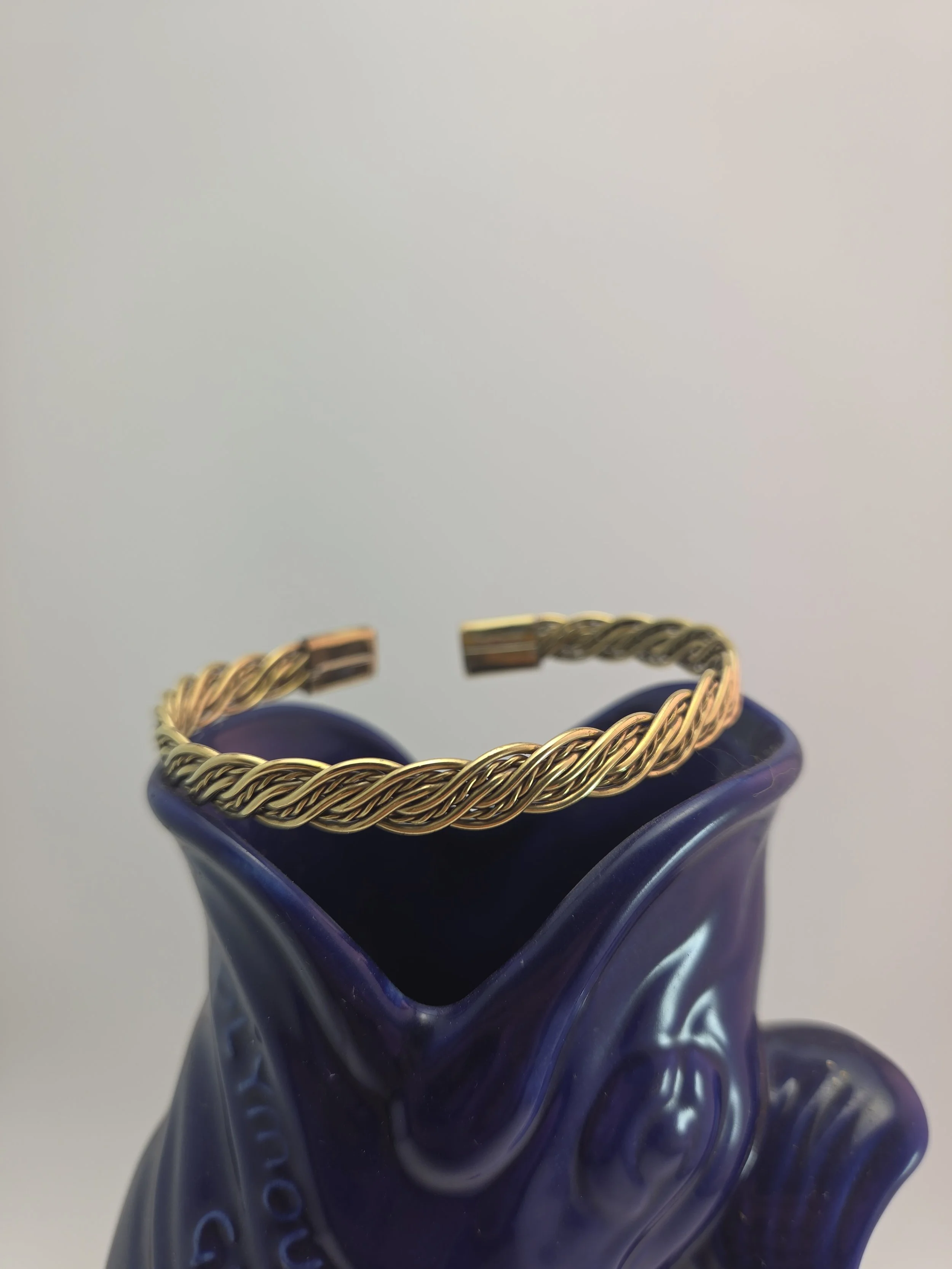 A brass chain bracelet with twisted links resting on the rim of a deep blue glass vase with a wavy, textured surface.