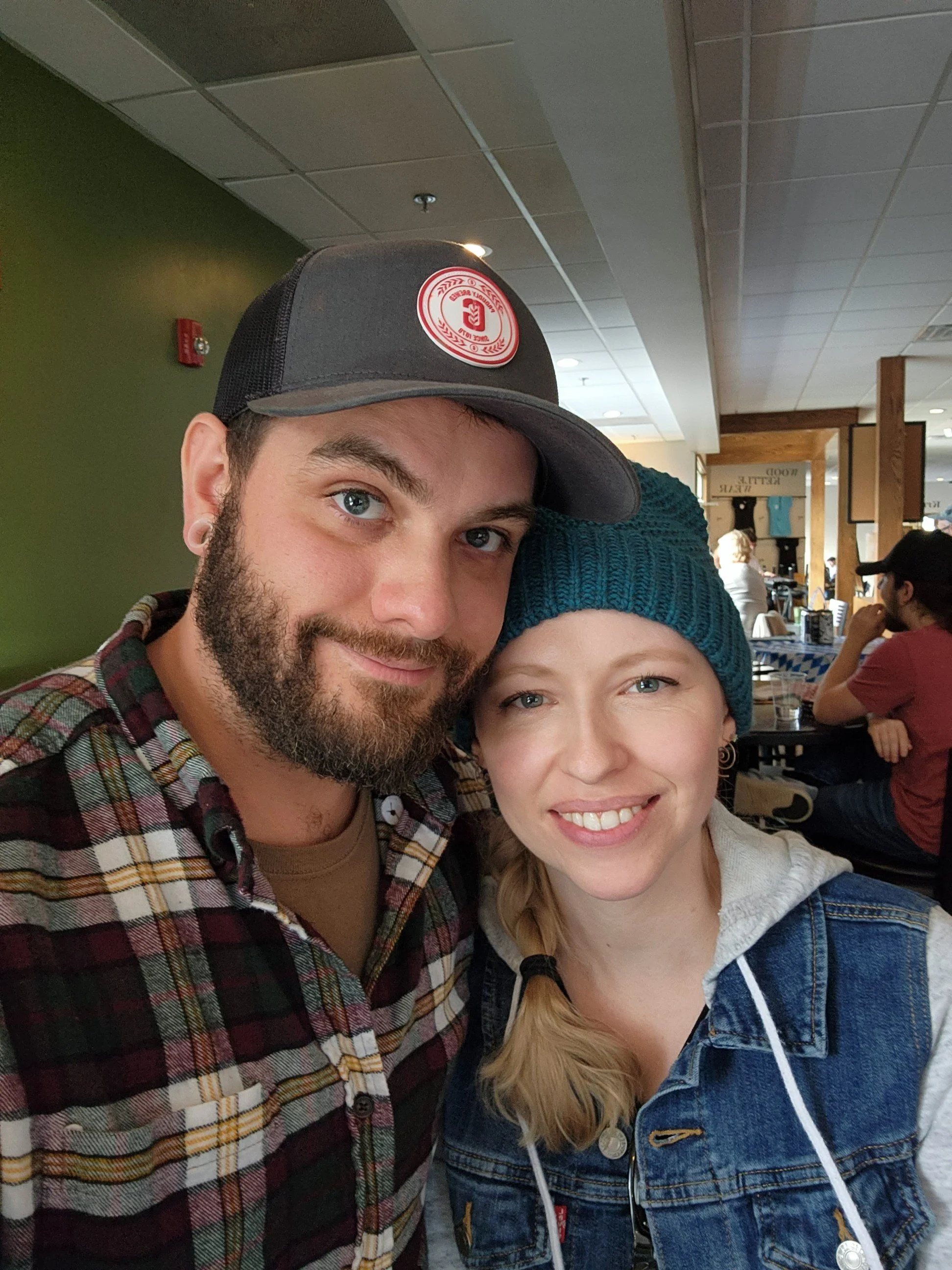 Close-up of two smiling people, a man with a beard wearing a baseball cap and a woman with blonde hair in a denim jacket, inside a cafe or restaurant.