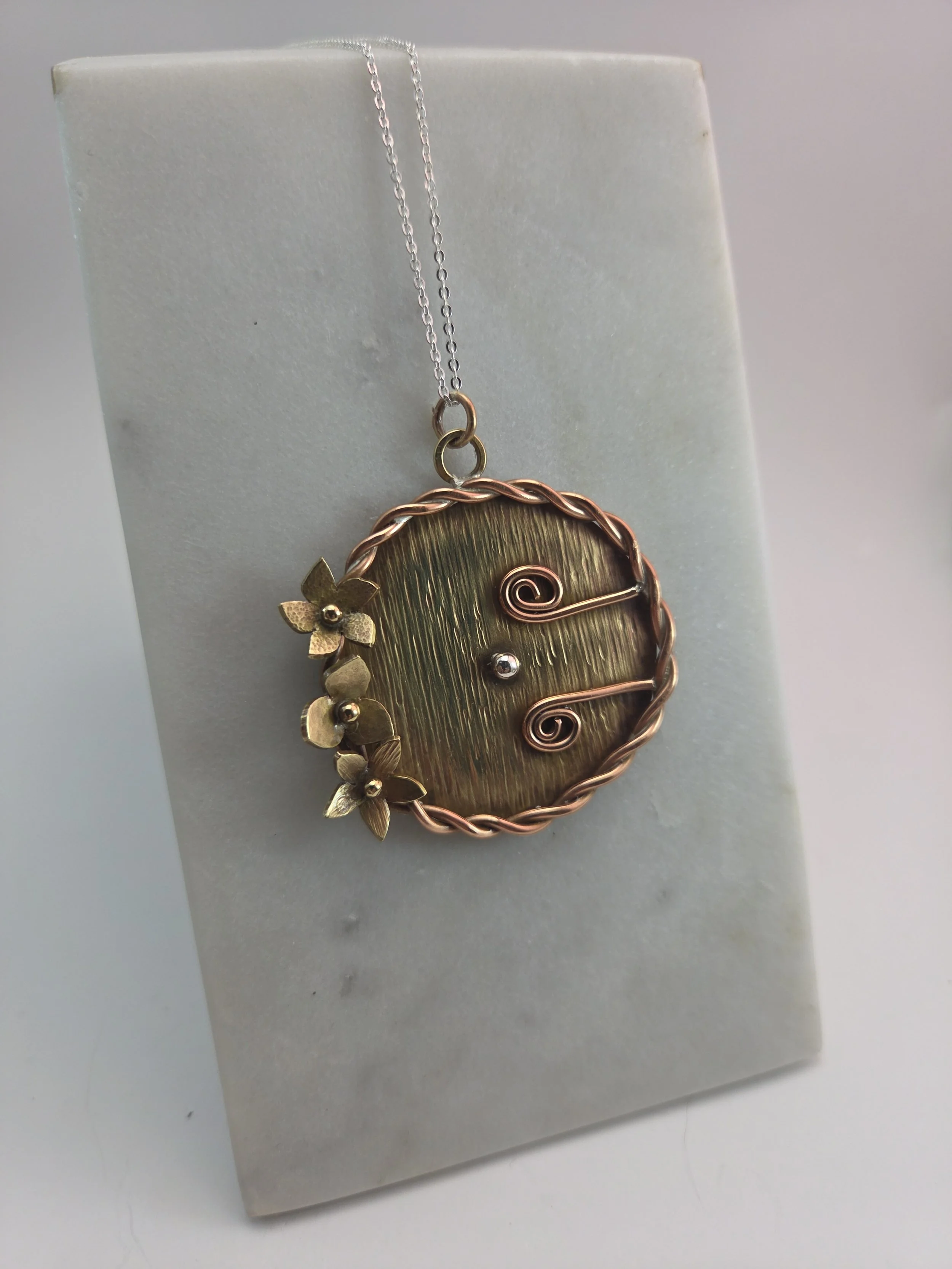 A decorative necklace with a large oval pendant featuring a textured brass and copper design, adorned with small brass flowers on the left side, hanging from a thin silver chain, displayed against a white background.