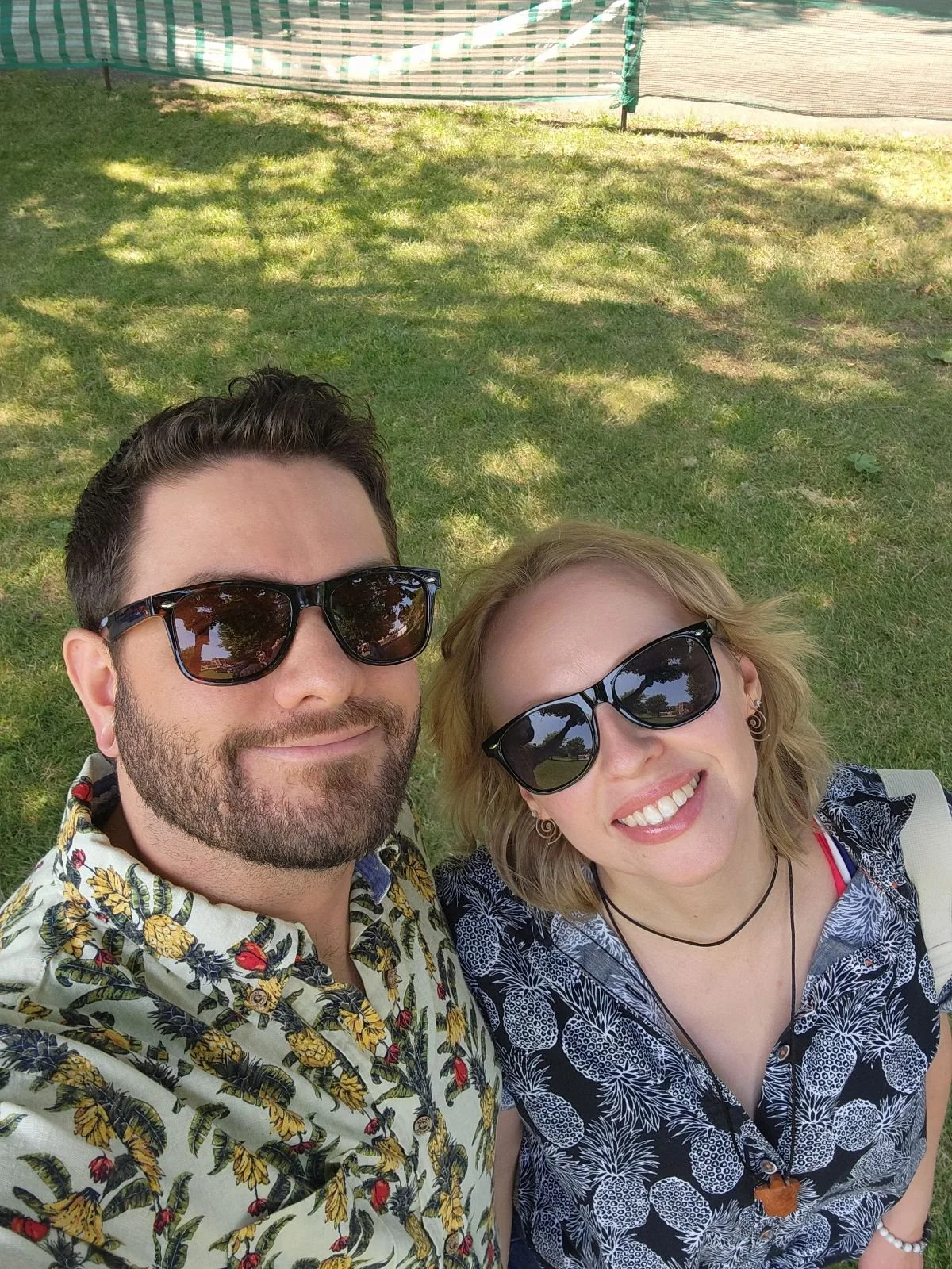 Two people taking a selfie outdoors on a sunny day, both wearing sunglasses and smiling, with a grassy area and a shaded background.