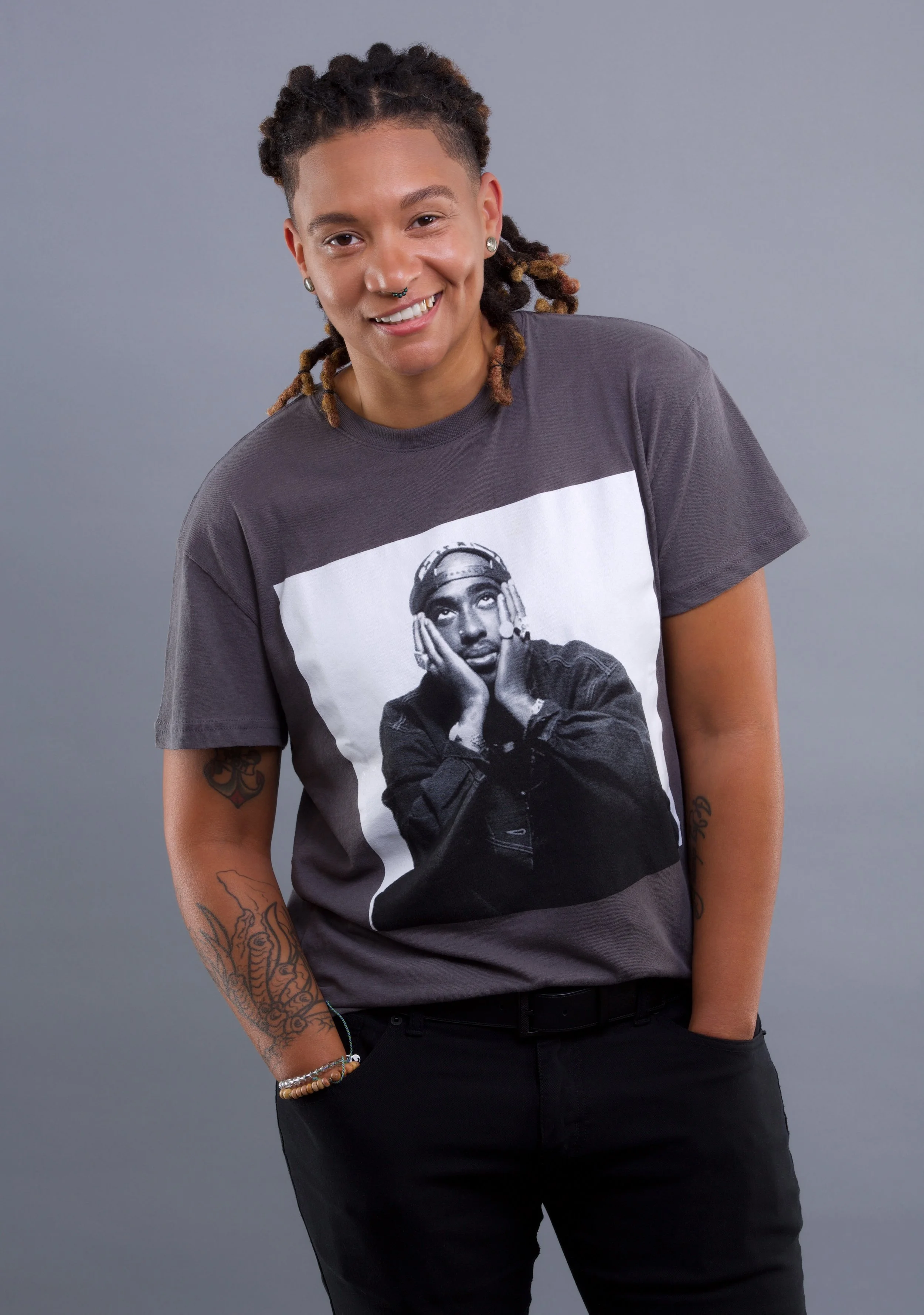 A person with dreadlocks and tattoos wearing a gray T-shirt featuring a black-and-white graphic of Tupac smiling against a plain gray background.