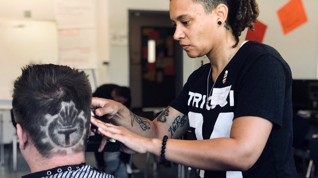 Dee Boozer as a barber with tattoos and black T-shirt giving a haircut to a man with a pattern shaved into his hair.