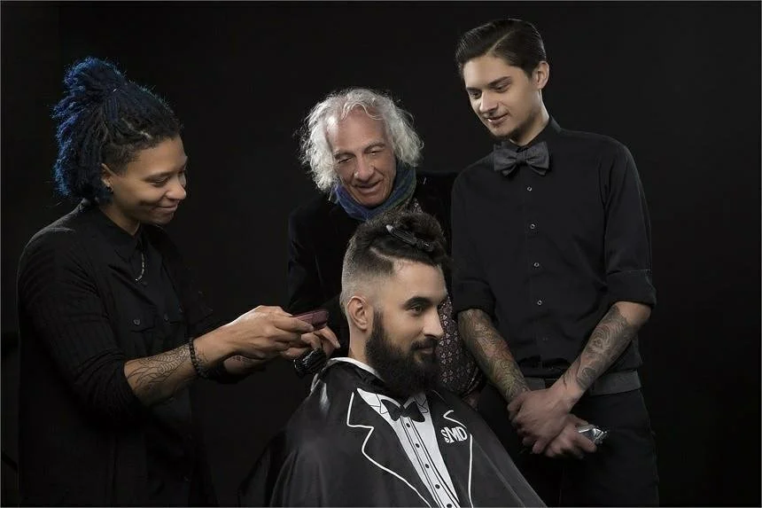 Dee Boozer giving haircut to a man with a beard, wearing a tuxedo cape, with Mario Tricoci and a young apprentice observing on a black background.