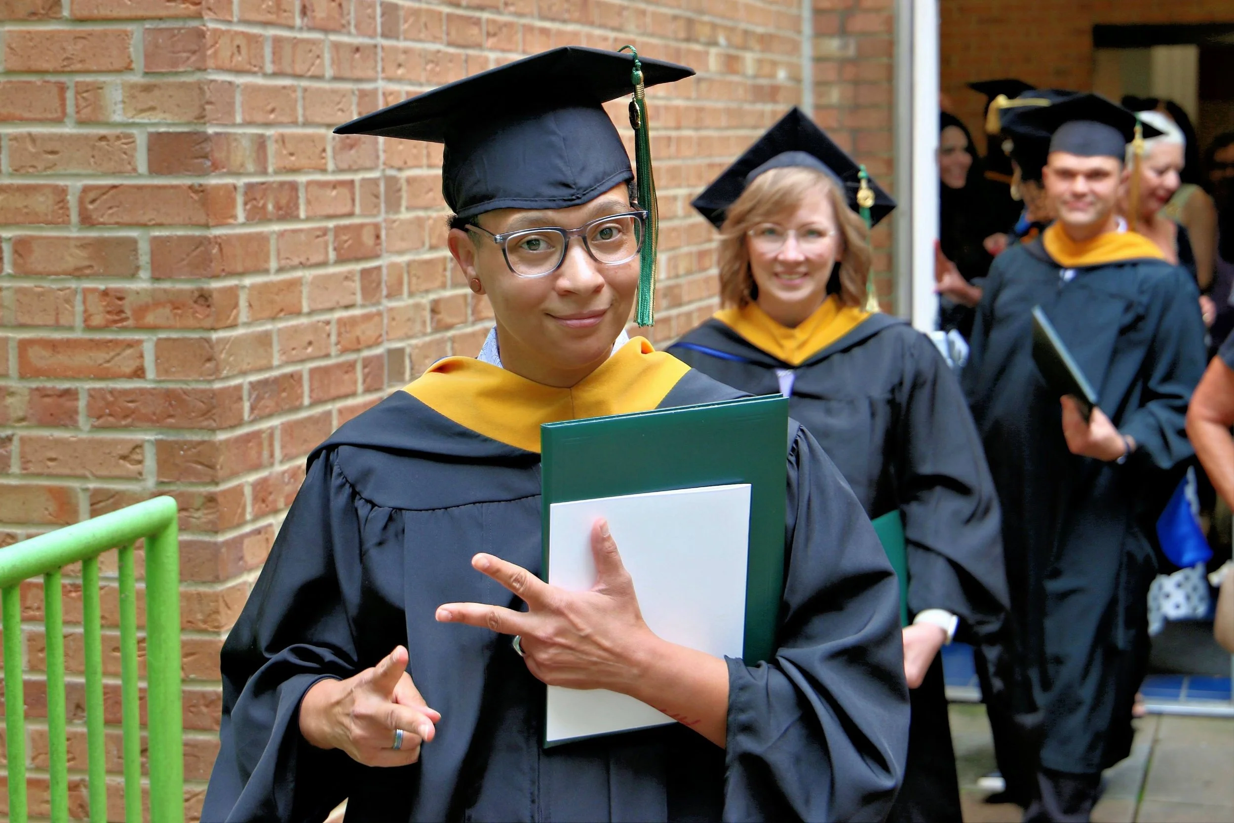Graduate woman wearing glasses and a black cap and gown holding a diploma, with graduation ceremony students in the background near a brick wall.