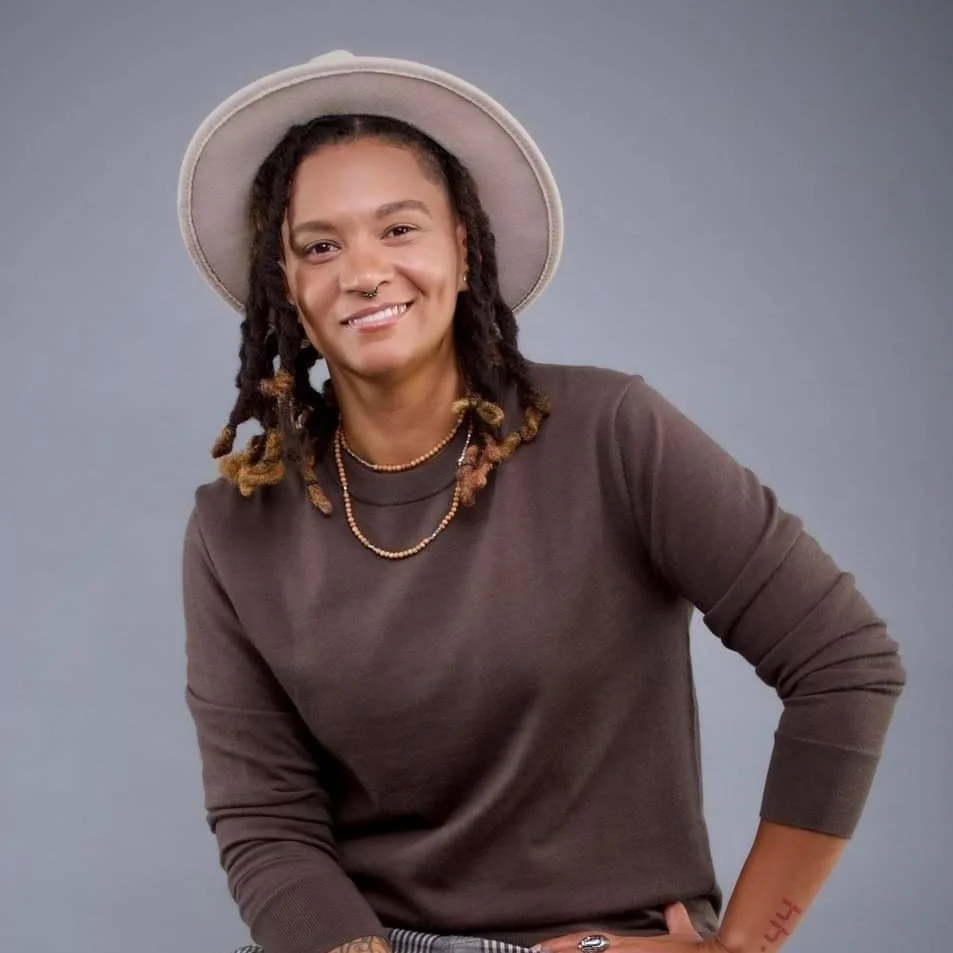 A woman wearing a beige hat, a brown long-sleeve shirt, and layered gold necklaces, smiling with dreadlocks and tattoos visible on her arm, standing against a plain gray background.