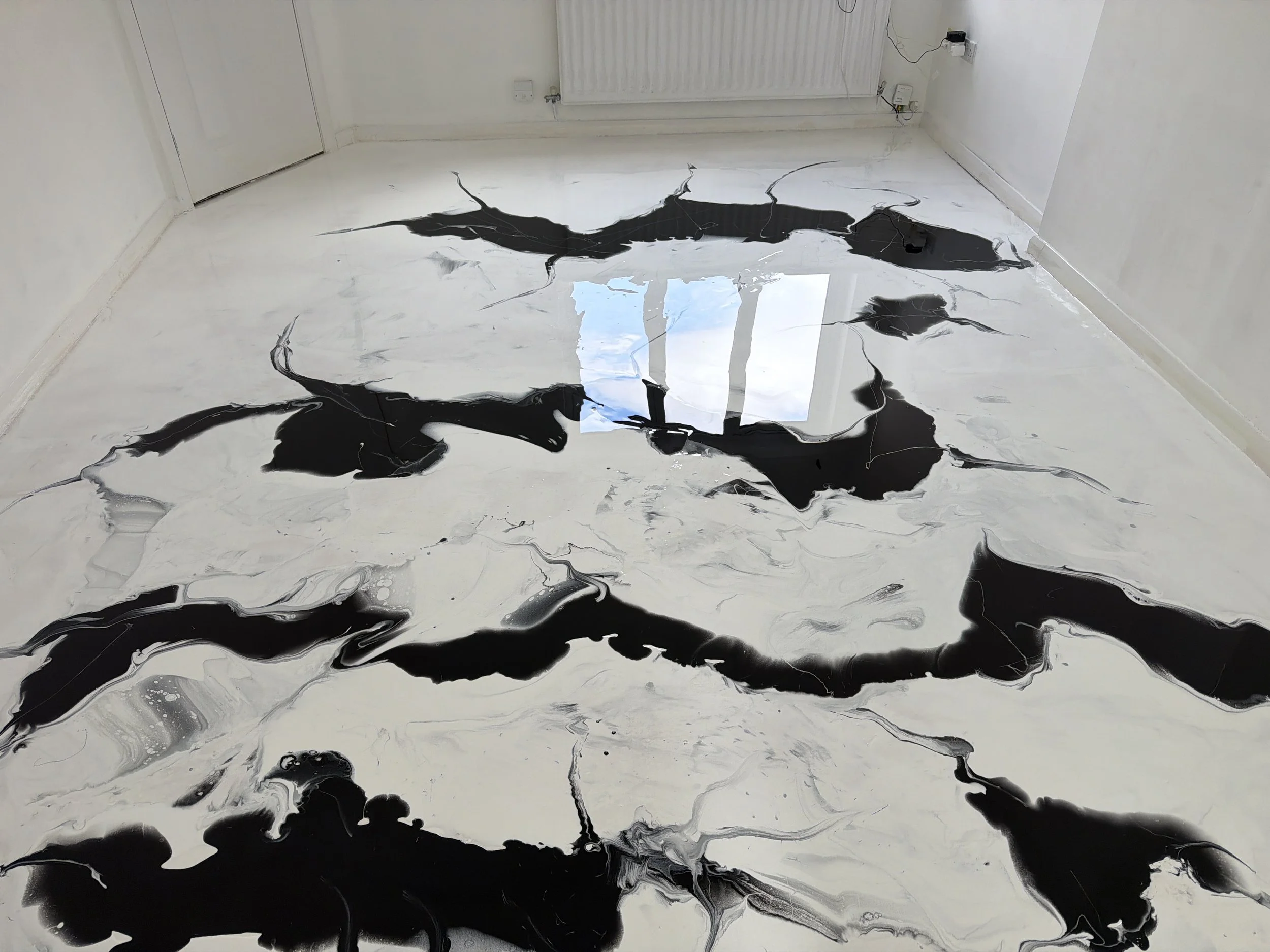 An abstract painting with black, white, and gray swirling patterns on a reflective floor in a white-walled room.