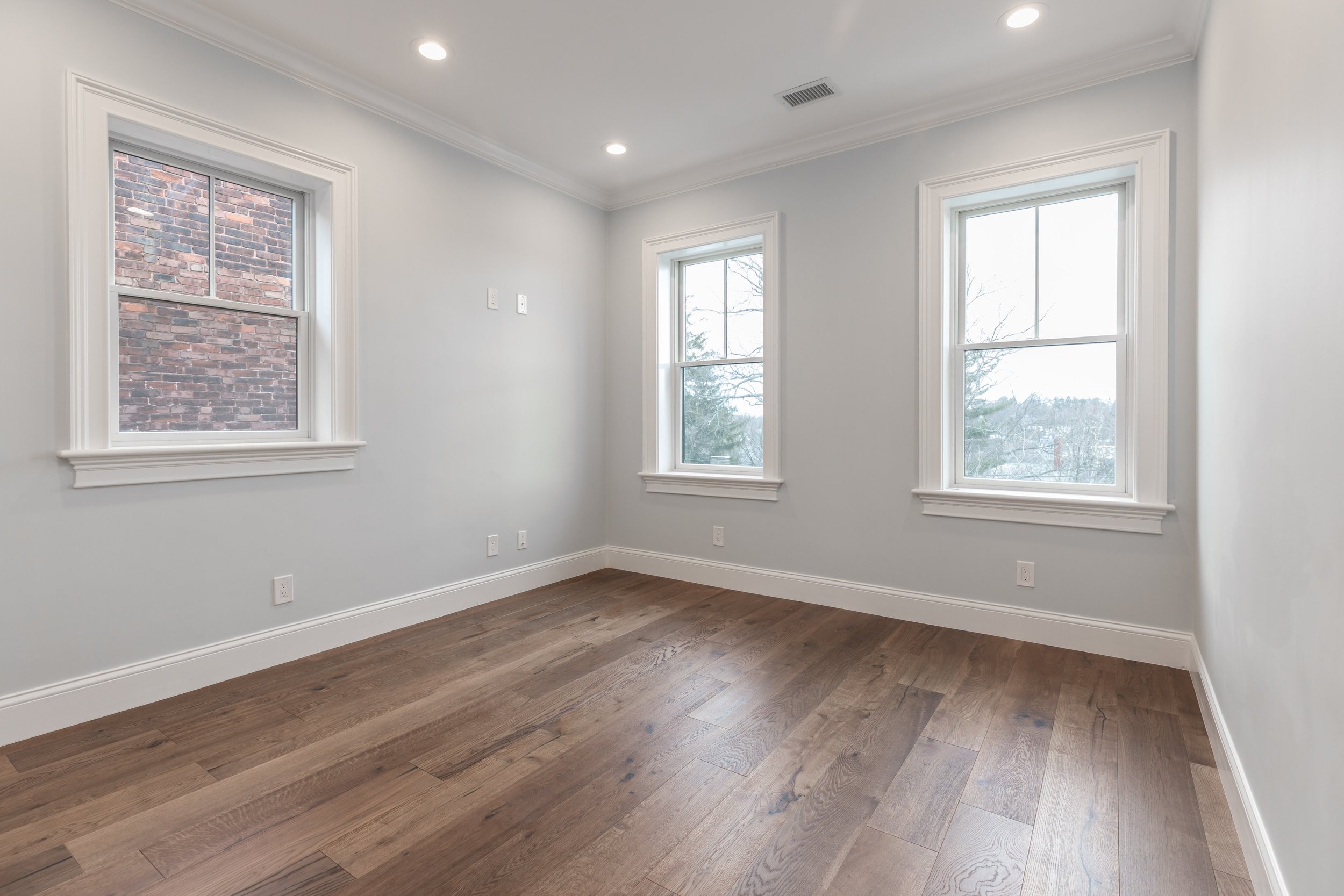 Empty room with three windows, white walls, hardwood flooring, ceiling lights, and electrical outlets.