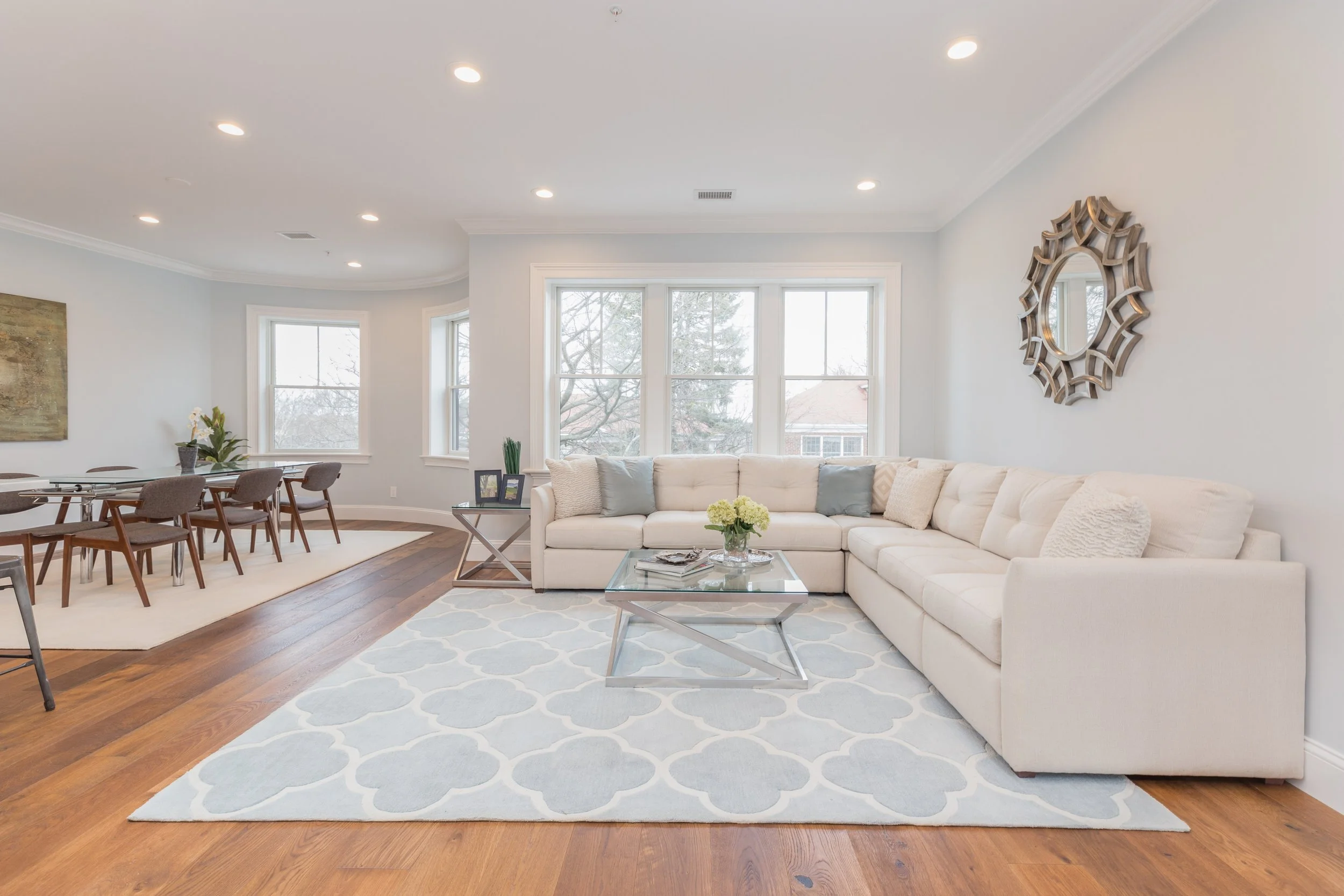 A spacious living room with a large cream sectional sofa, a glass coffee table, and a patterned area rug. There are multiple large windows with a view of trees outside. The room is decorated with wall art, a circular mirror, and some framed photograp