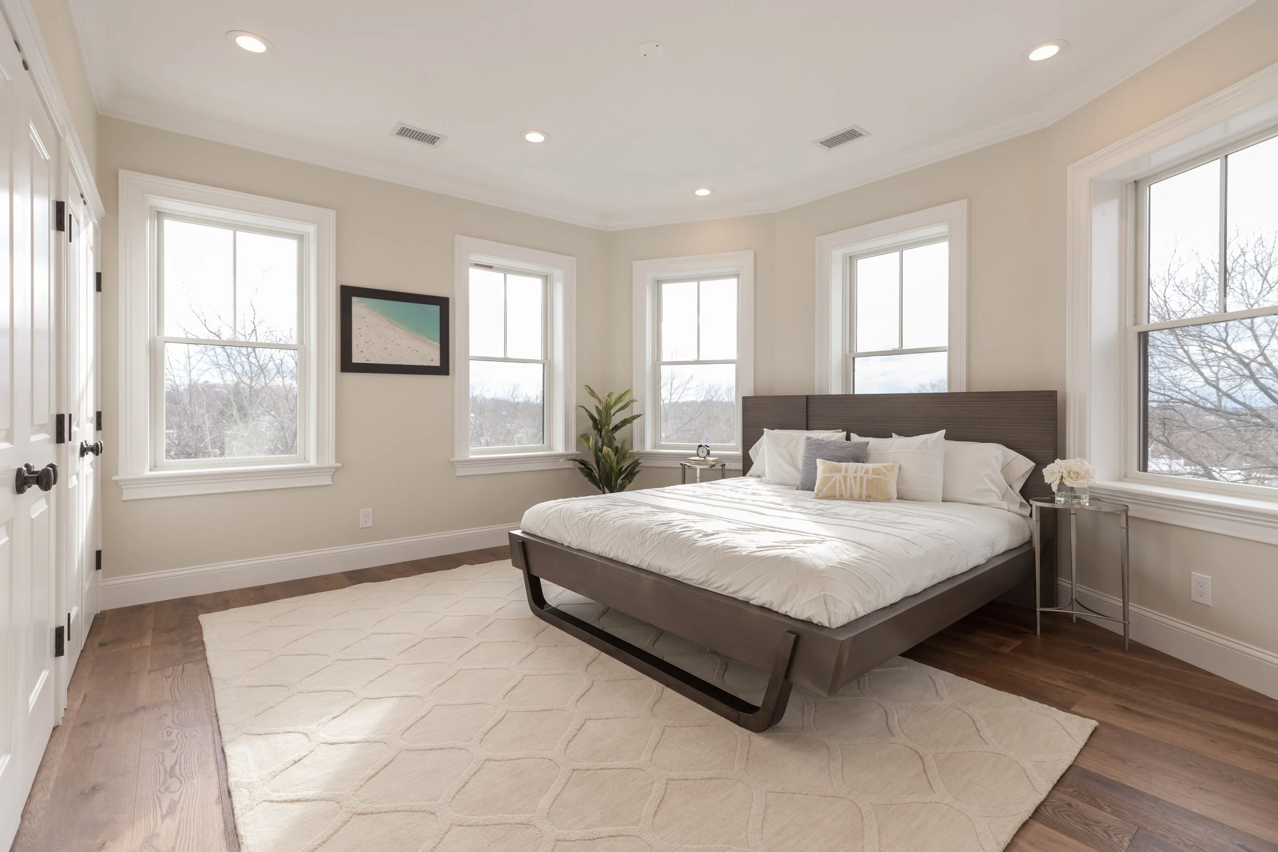 A bedroom with a large bed, four windows, a plant, a framed beach picture, and a white rug on wooden flooring.
