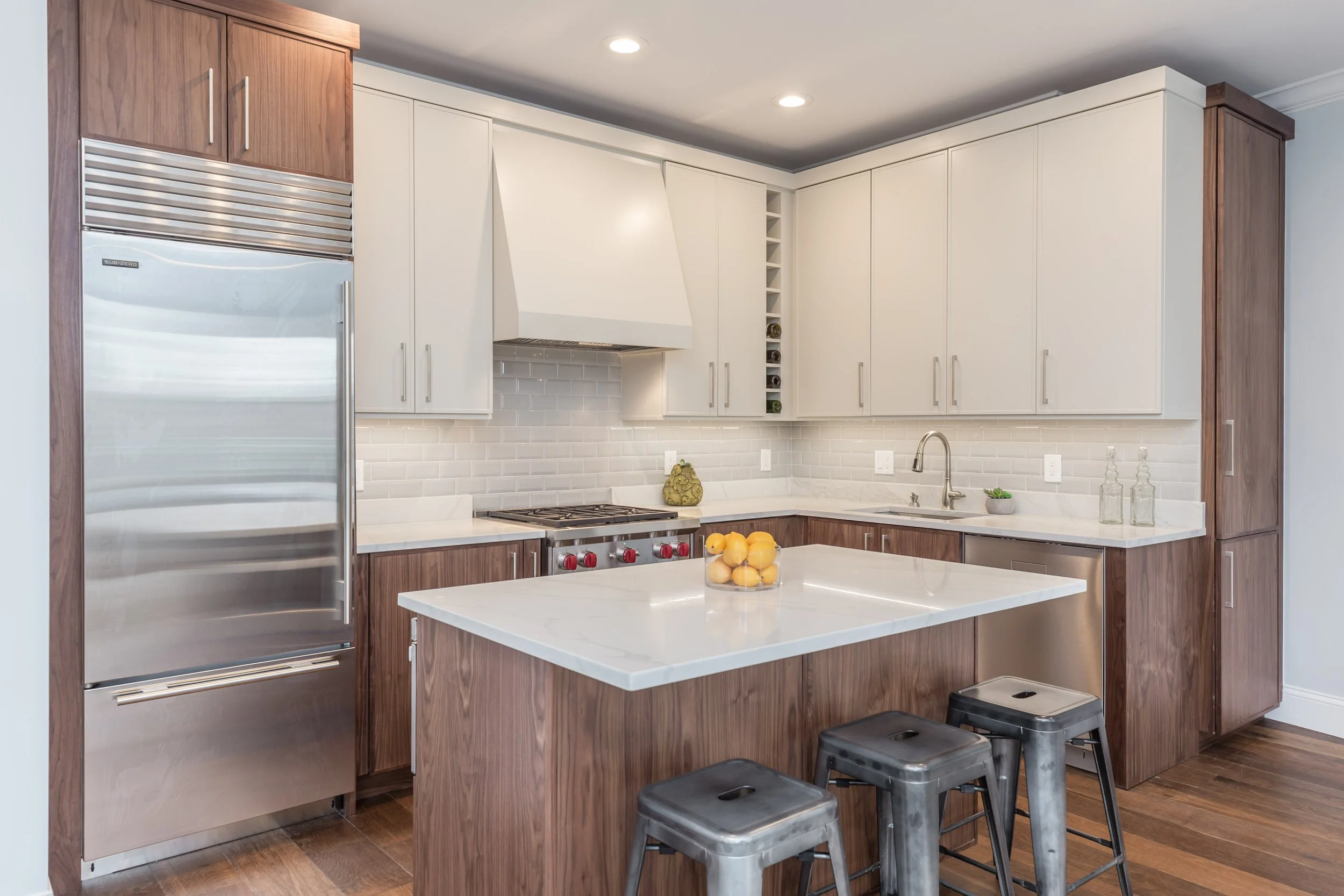 Modern kitchen with white cabinetry, wooden accents, stainless steel appliances, and a central white marble island with lemons, two black stools, and a sink.