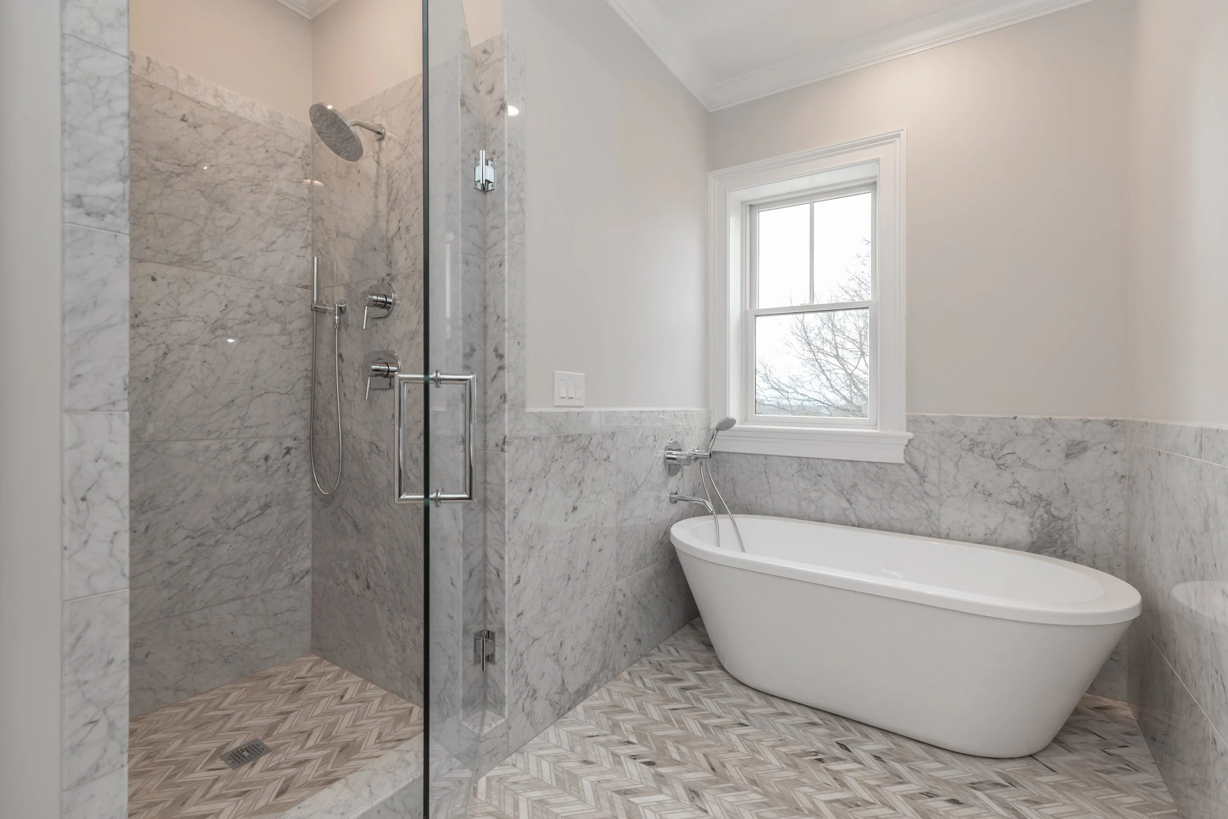 Modern bathroom with marble walls, a shower area with glass door and a rainfall showerhead, and a freestanding bathtub next to a window.