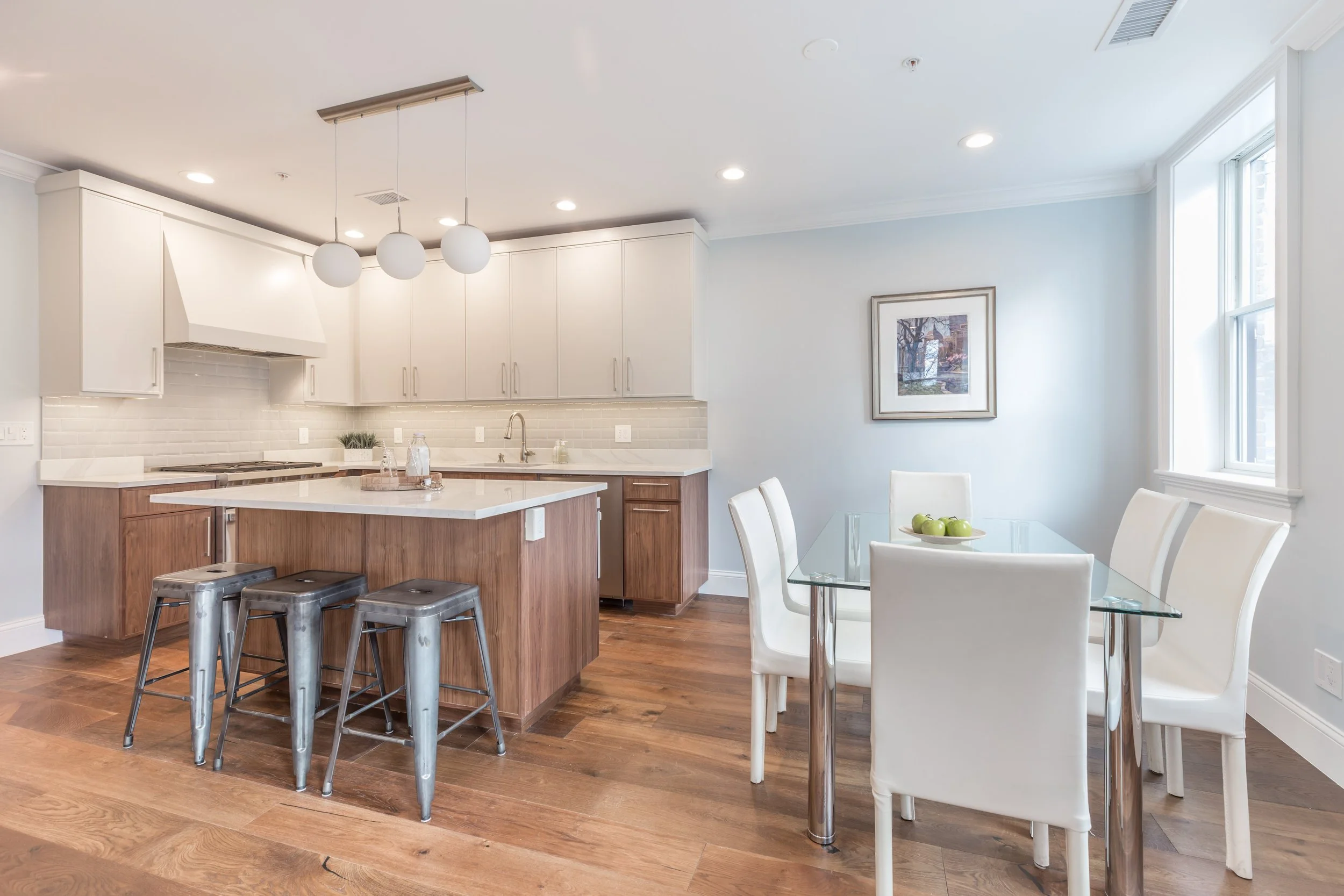 Bright modern kitchen with white cabinets, a kitchen island with a marble countertop, stainless steel stools, a dining area with a glass table and white chairs, a window with sunlight, framed artwork on the wall, and hardwood flooring.