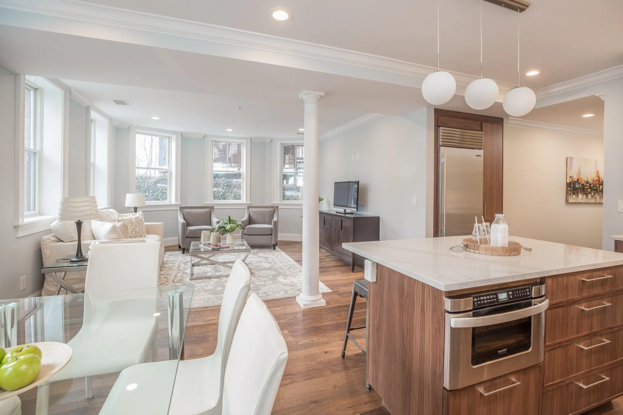 Open-concept living room and kitchen with white walls, hardwood floors, large windows, white sofa, gray armchairs, flat-screen TV, modern pendant lights, marble kitchen island, stainless steel appliances, and minimalistic decor.