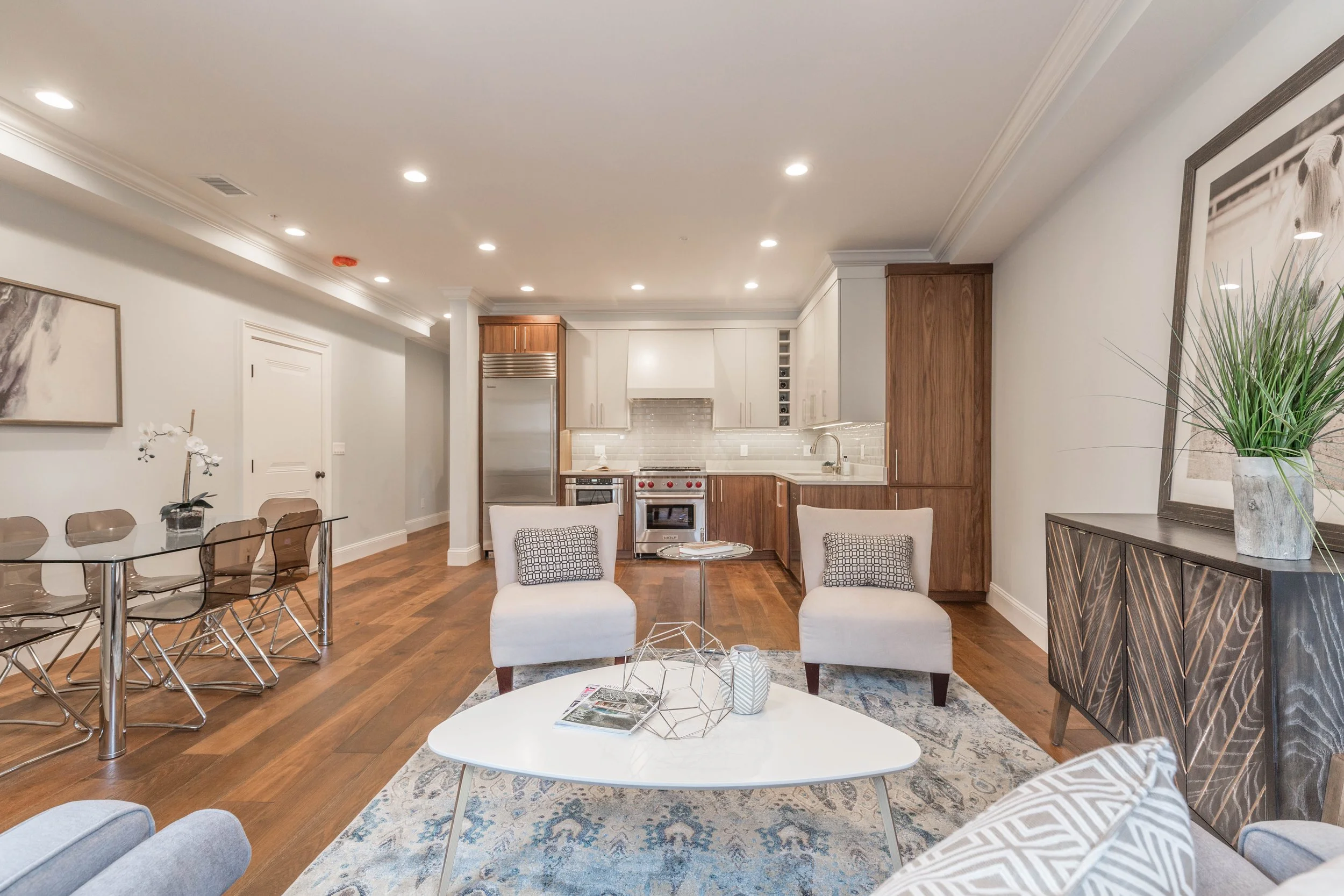 Open-concept living room and kitchen with dining area, modern furniture, hardwood floors, white walls, recessed lighting, and decorative plants.