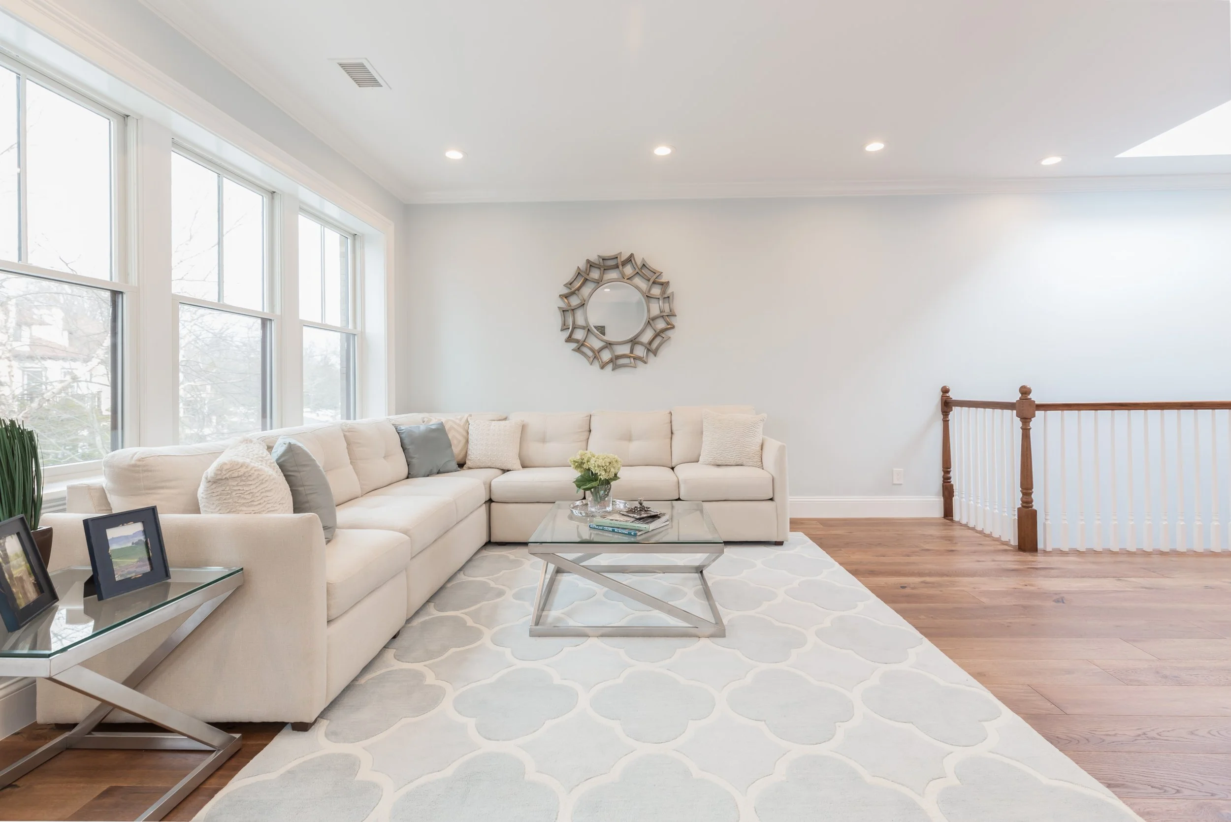 Living room with large window, white sectional sofa with pillows, glass coffee table with books and flowers, decorative mirror on wall, hardwood floor, and staircase railing.