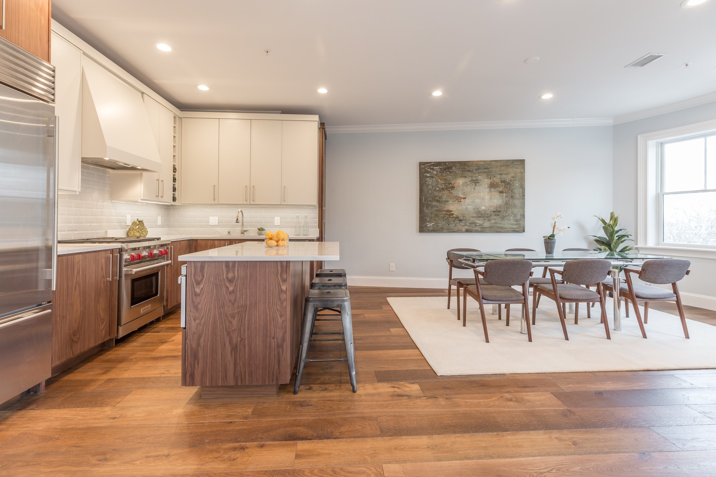 Open concept kitchen and dining area with wooden flooring, white upper cabinets, wooden lower cabinets, a kitchen island with bar stools, and a dining table with chairs. There is a large window, a piece of abstract wall art, and a plant.
