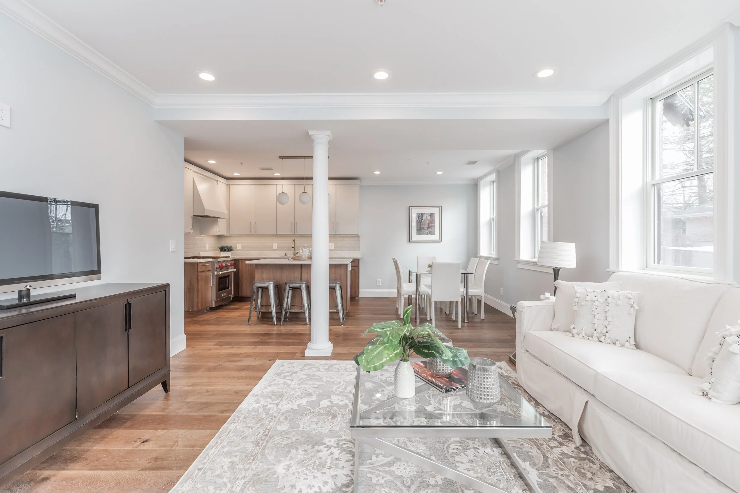 Living room and kitchen area with white walls, wooden flooring, and large windows. The living room has a white sofa with decorative pillows, a glass coffee table with plants and books, and a patterned rug. The kitchen features white cabinets, a count