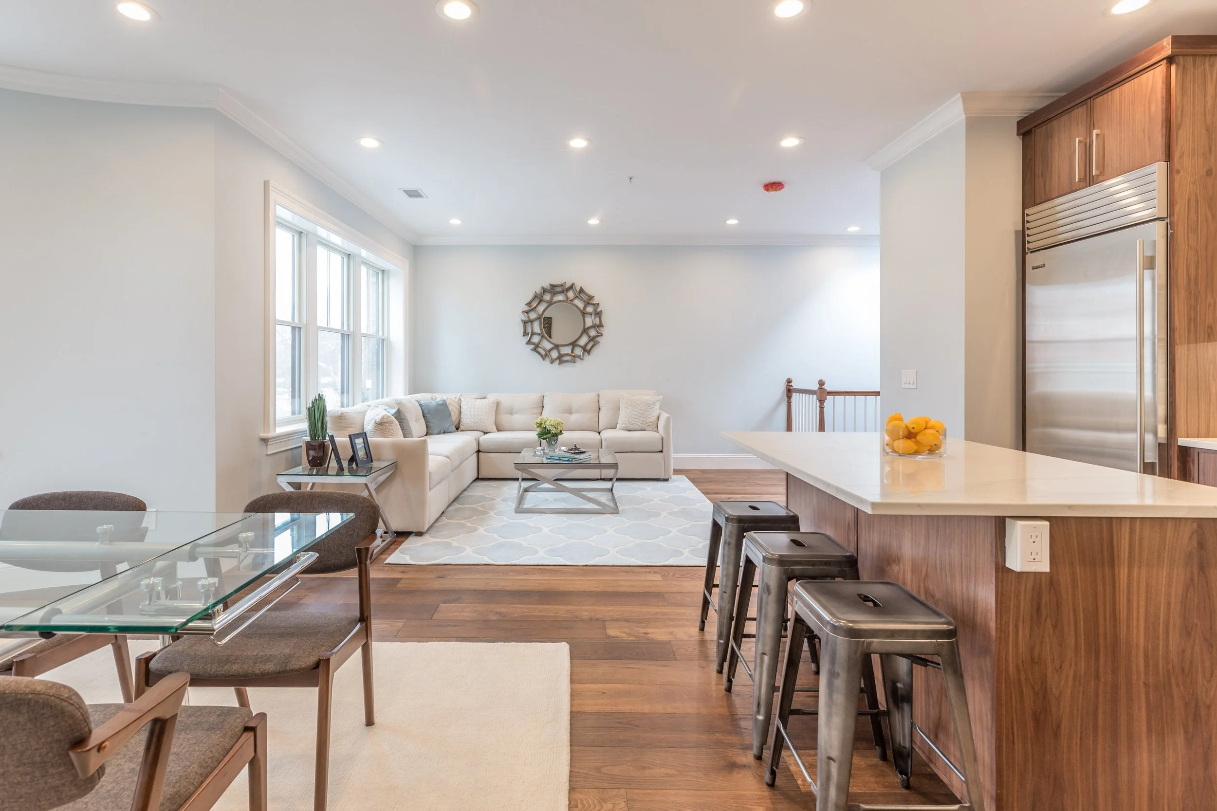 Open-concept living room and kitchen with large windows, beige sofa with pillows, wooden dining table with chairs, wooden kitchen cabinets, stainless steel refrigerator, and a glass bowl of oranges on the kitchen counter.