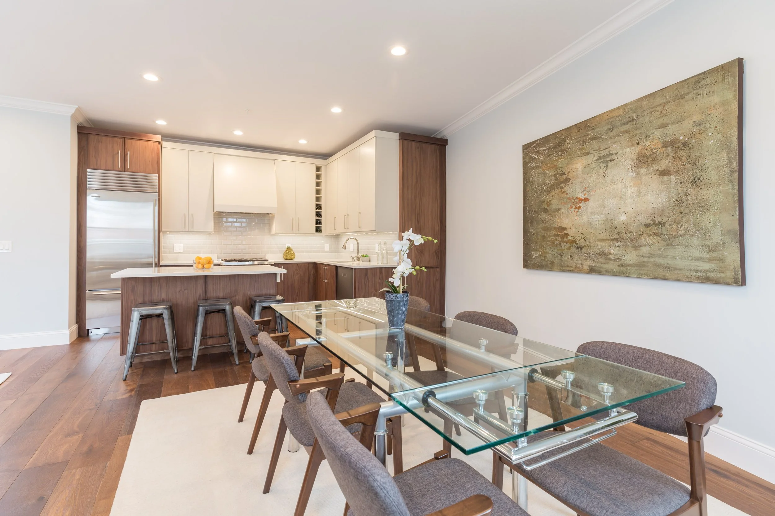 A modern kitchen and dining area with wooden flooring, white and wooden cabinets, a glass dining table with brown upholstered chairs, and a large abstract painting on the wall.