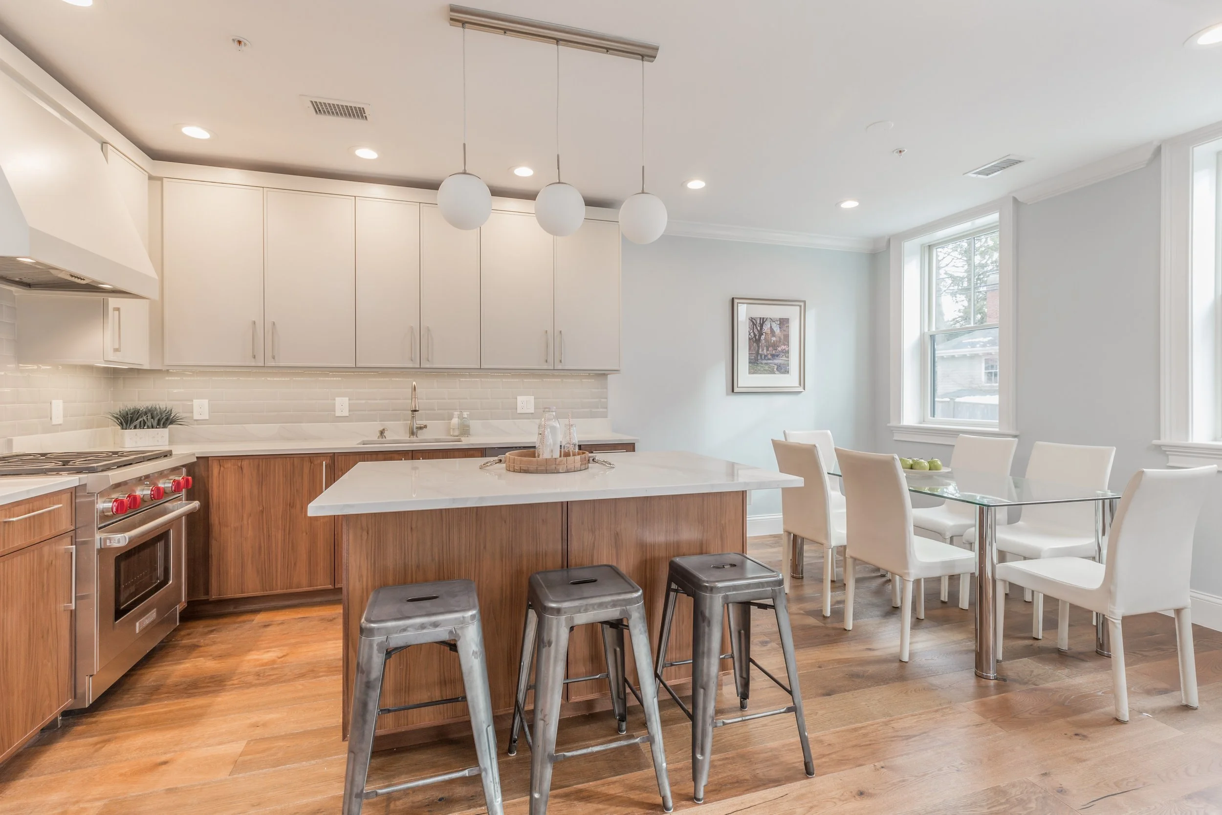 Modern kitchen with white cabinets, a white countertop island, stainless steel appliances, hanging white spherical pendant lights, and a dining area with white chairs and a glass table near large windows.
