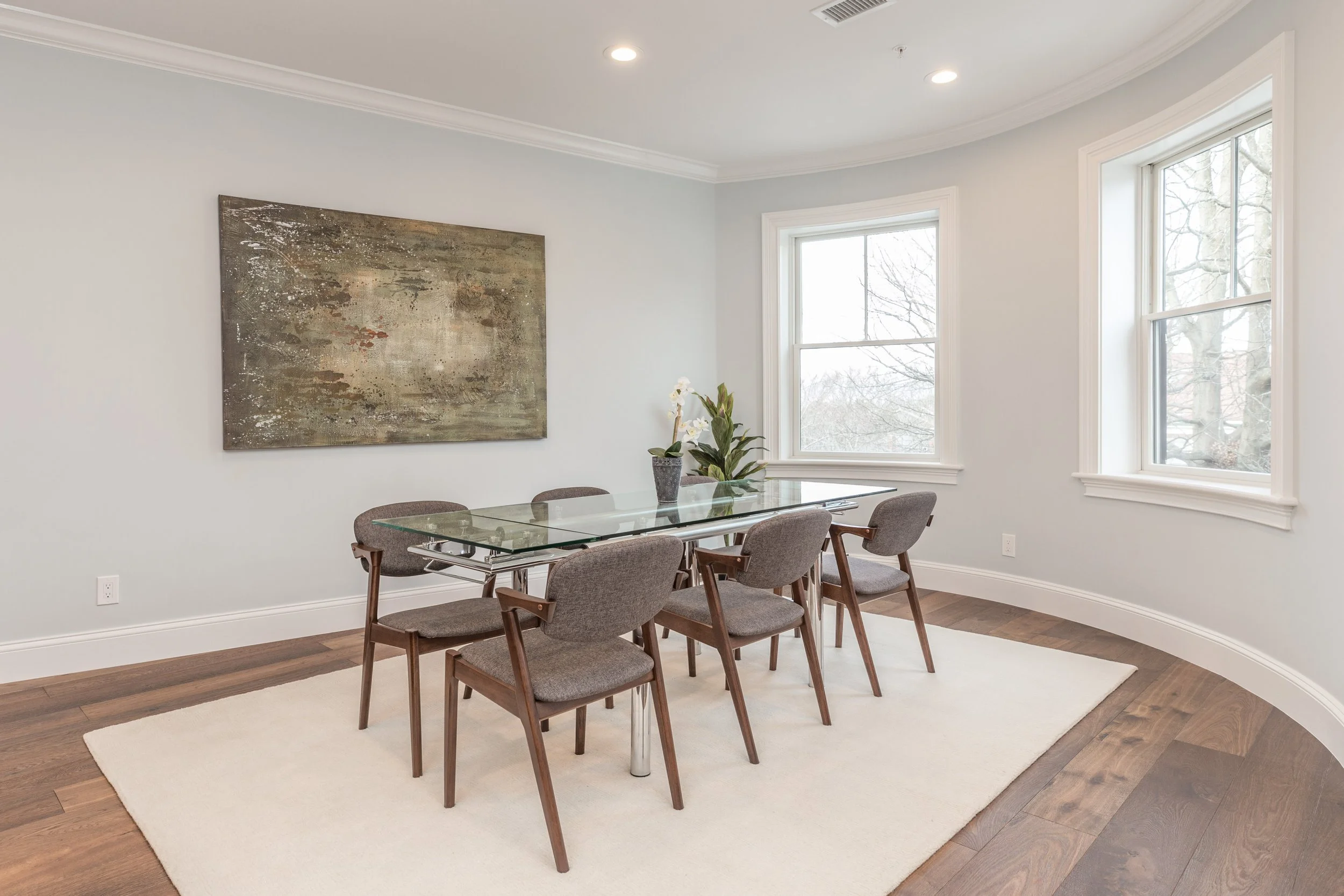 A dining room with a glass-top table surrounded by six brown upholstered chairs, a large abstract painting on the white wall, two windows providing natural light, a potted plant on the table, and hardwood flooring with a white rug.