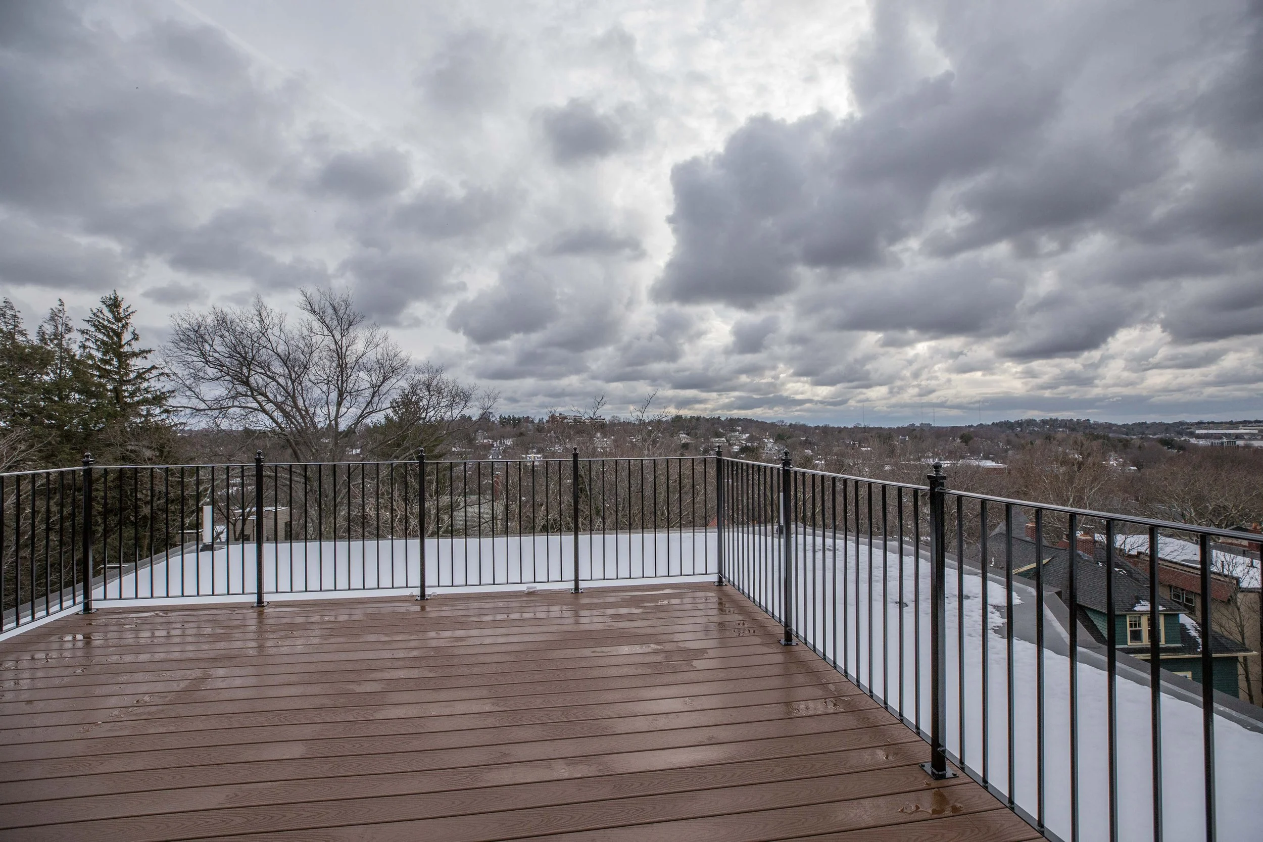 A view from a wooden balcony with a black metal railing, looking out over a snow-covered landscape with trees and rooftops under a cloudy sky.