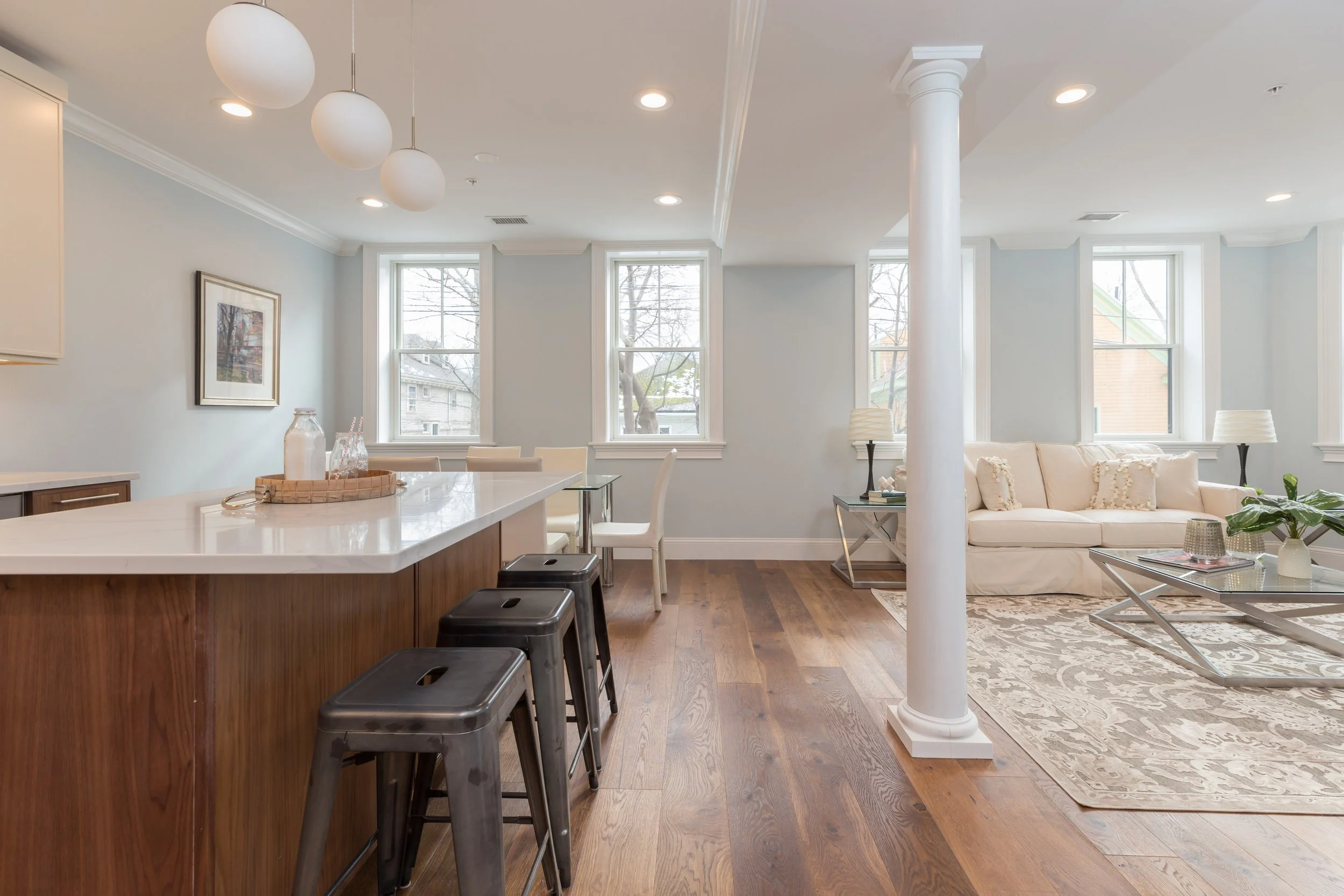 Bright living room with large windows, a white couch, a marble coffee table, a kitchen island with black stools, and hanging ceiling lights.