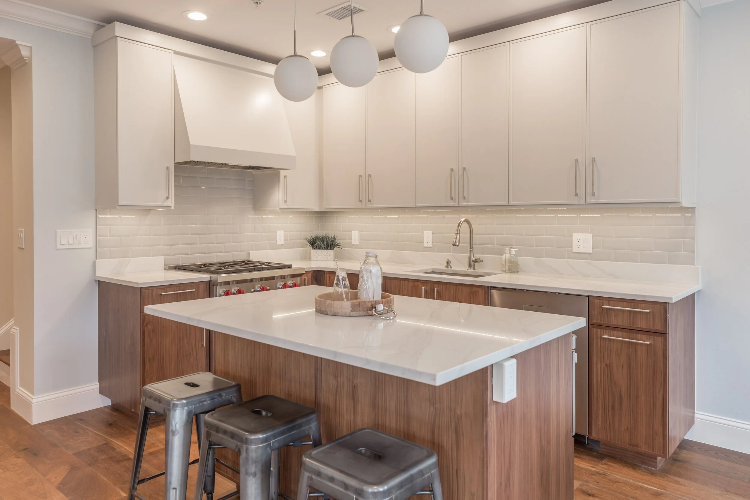 Modern kitchen with white upper cabinets, brown lower cabinets, white countertops, a stainless steel stove, a white subway tile backsplash, three white pendant lights, and a kitchen island with metal stools.