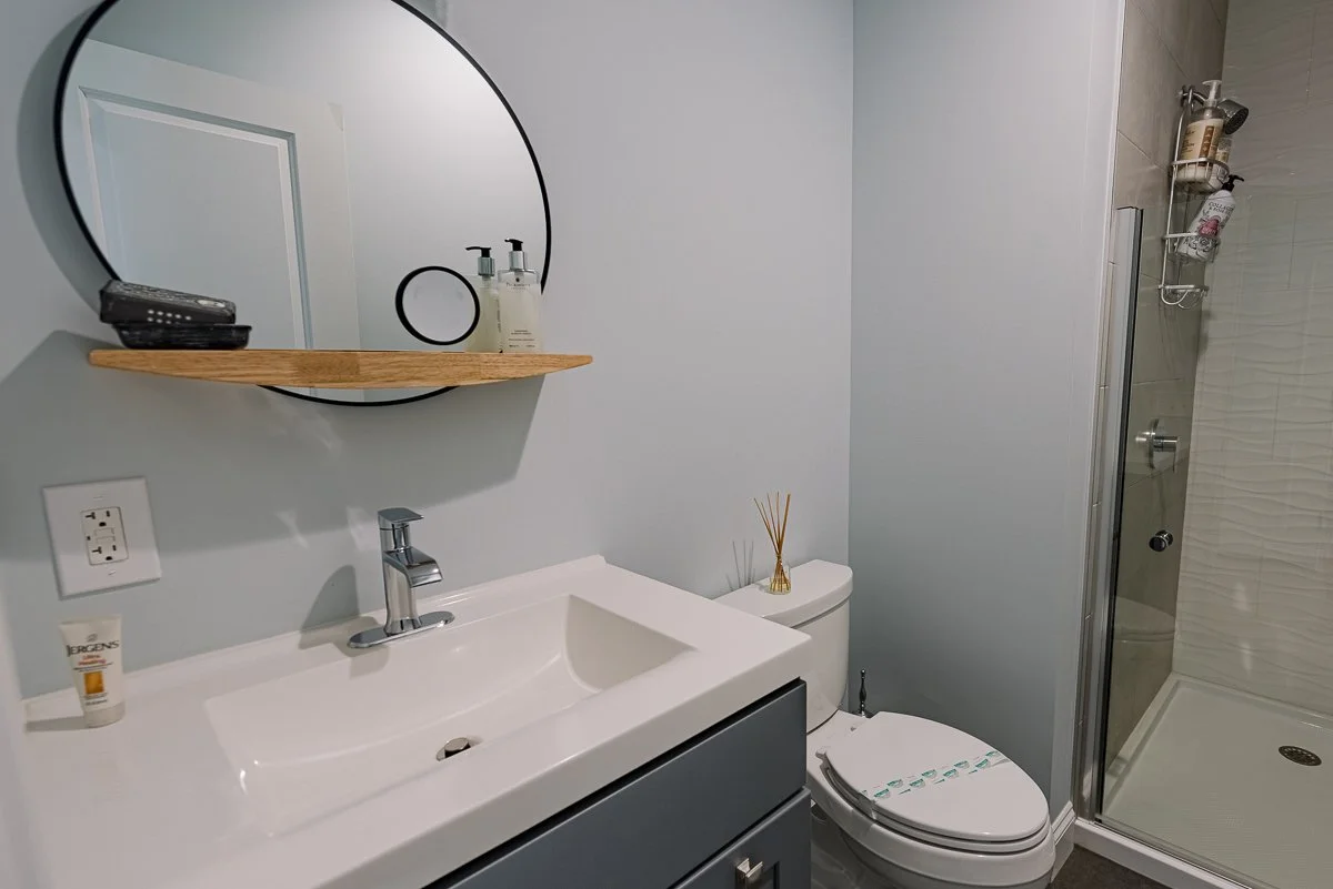 Bathroom with white sink, navy vanity, circular mirror, small shelf with soap dispenser and remote, toilet with a decorative reed diffuser, and a glass shower enclosure with shower products inside.