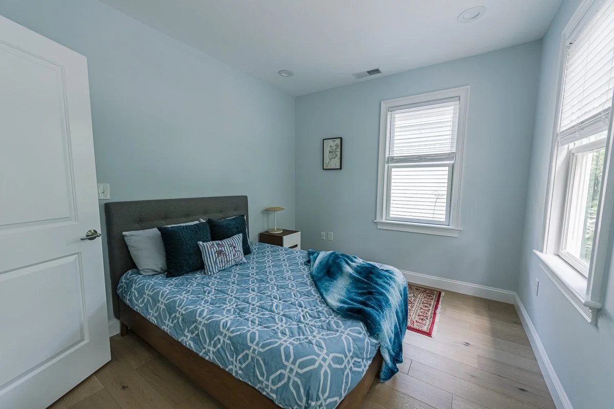 A bedroom with a bed having a dark gray upholstered headboard, blue patterned bedding, and several pillows. There are two windows with white blinds, a nightstand with a small lamp, a framed picture on the wall, a small red rug, and light-colored wood