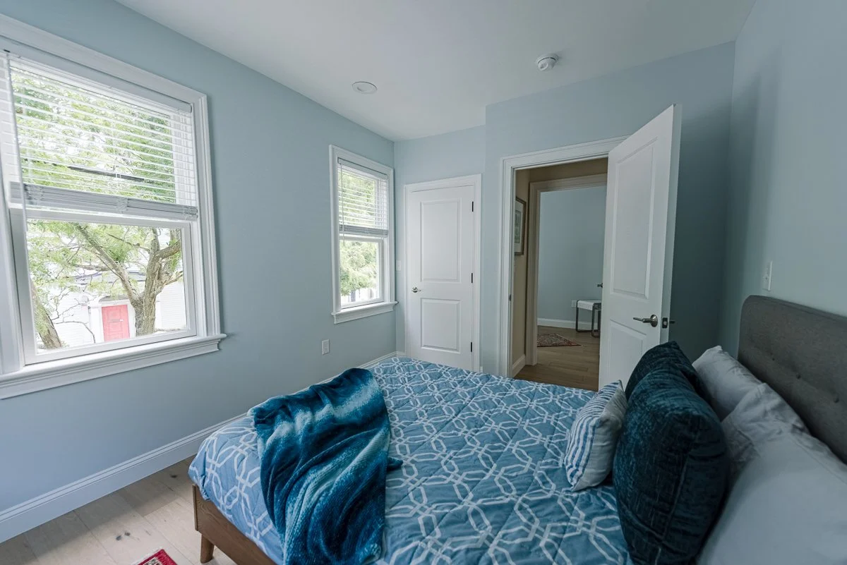 Bedroom with light blue walls, two windows, a bed with blue and gray bedding, and an open door leading to another room.
