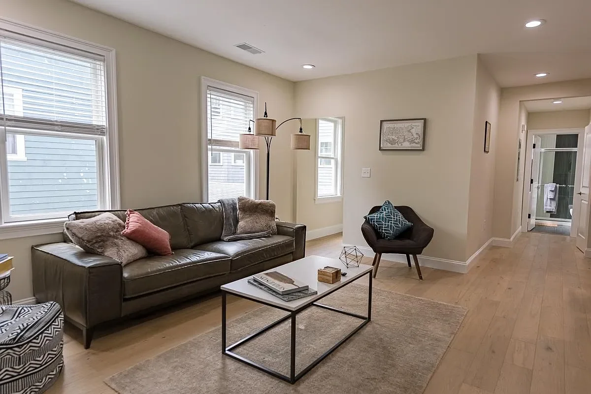 Living room with a brown leather sofa, decorative pillows, a black armchair with a blue pillow, a white coffee table, and a beige area rug, with multiple windows and hallway leading to a bathroom.