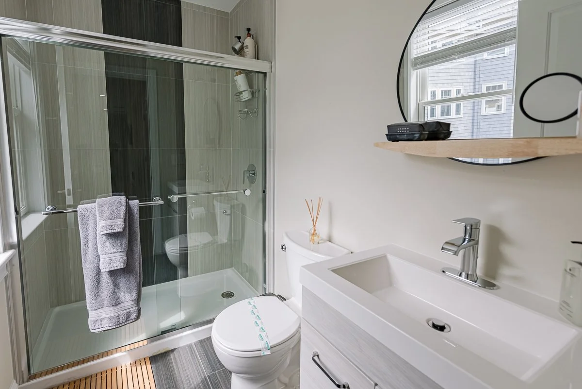 Bathroom with a glass shower, white toilet, white sink, a round mirror, and a window with blinds.