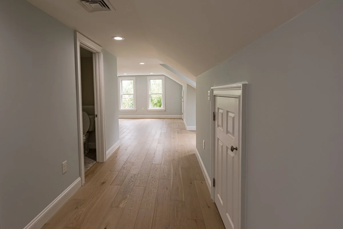 Empty room with hardwood floors, light blue walls, and three windows at the far end letting in natural light. There is a small, white door on the right wall and an open doorway leading to a bathroom with a visible toilet on the left.