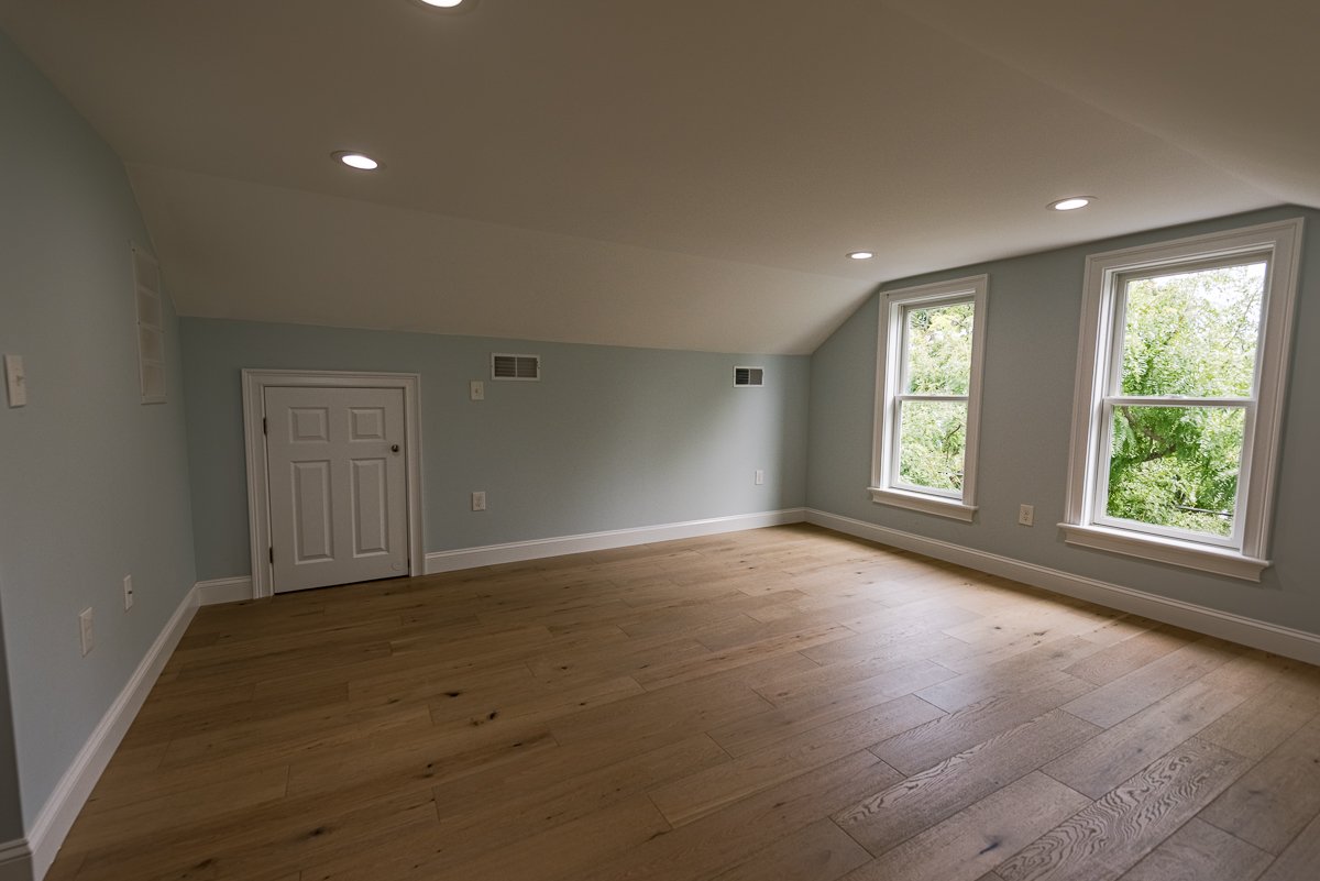 Empty upstairs room with light blue walls, wooden flooring, three windows showing greenery outside, and small white door on wall.