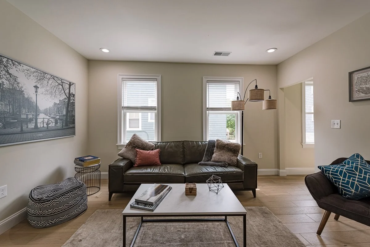 Living room with a dark leather sofa, two windows with blinds, a coffee table, decorative pillows, wall art, a standing lamp, and a ceiling light fixture.