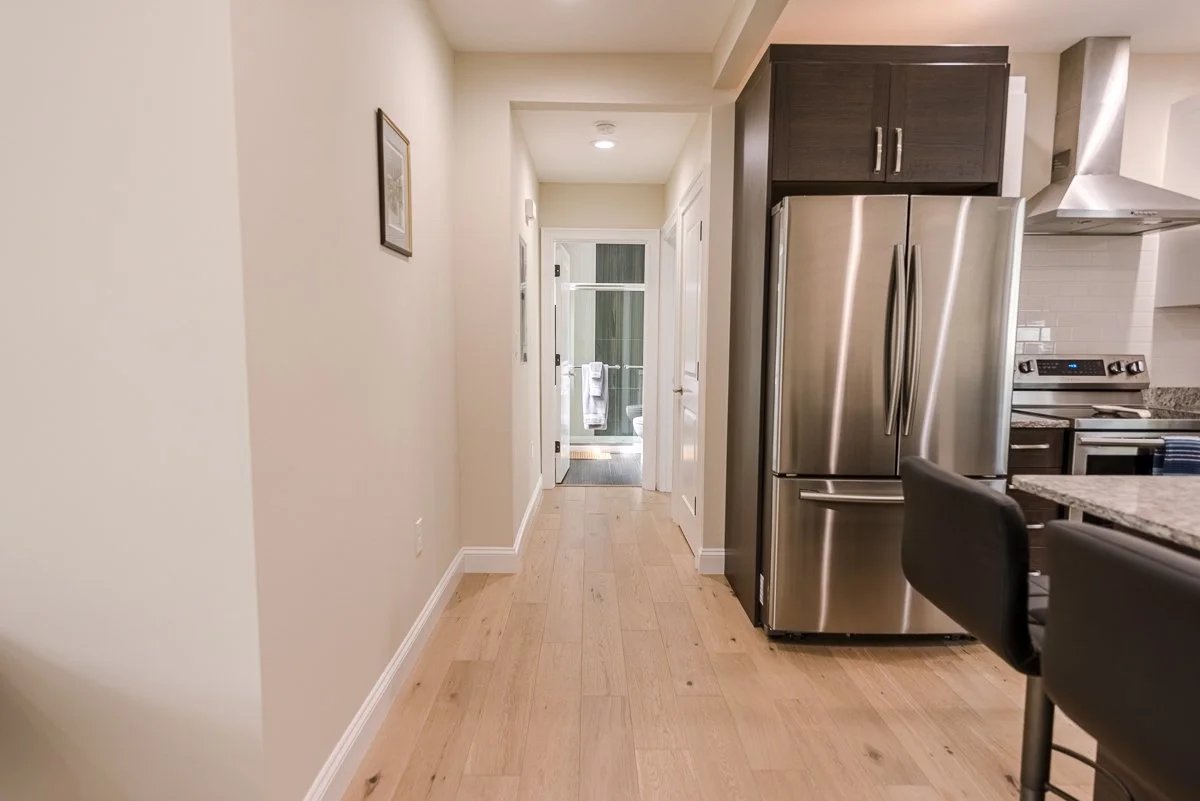 A kitchen view showing a stainless steel refrigerator, dark cabinetry, a stove, and a vent hood. A hallway with a wooden floor leads to a door with a glass window, revealing an outdoor area with a towel hanging.