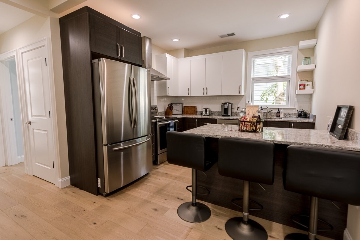 Modern kitchen with stainless steel refrigerator and stove, white and dark wood cabinets, granite countertops, and a breakfast bar with three black barstools.