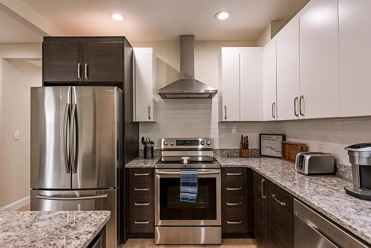Modern kitchen with stainless steel refrigerator, oven, and range hood, white and dark wood cabinets, granite countertops, and small appliances.