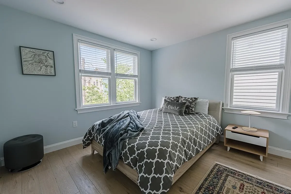 A bedroom with light blue walls, two windows with white blinds, a bed with a patterned gray and white comforter, and pillows, one with the word 'home'. There is a small nightstand with a lamp and remote, a black ottoman, and part of a tapestry rug vi