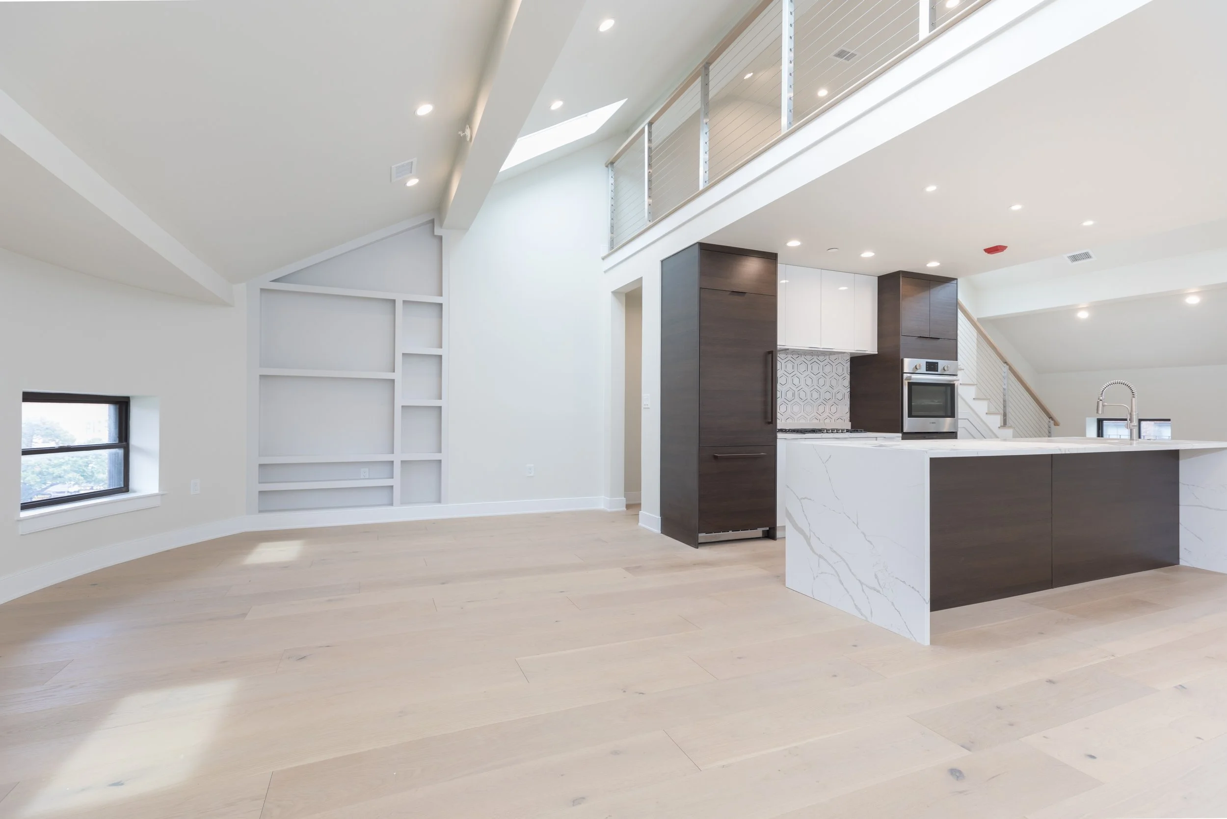 Modern open-plan kitchen with white and dark wood cabinetry, marble island, stainless steel appliances, high ceilings, and large windows with natural light.